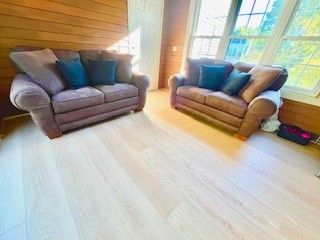 Two matching brown loveseats with blue accent pillows sit on light-colored wood flooring near a window.
