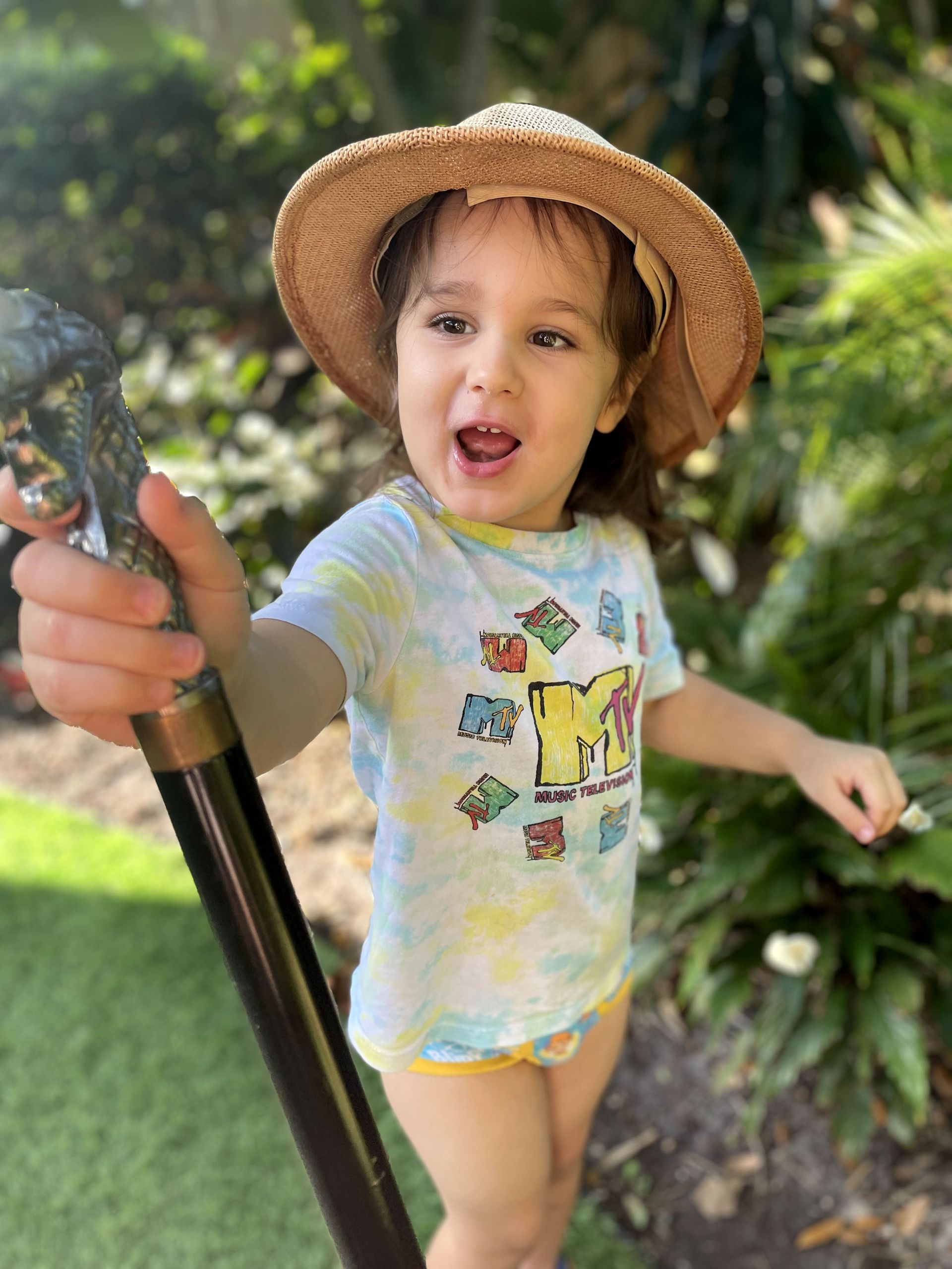 A little girl wearing a straw hat and a tie dye shirt is holding a cane.