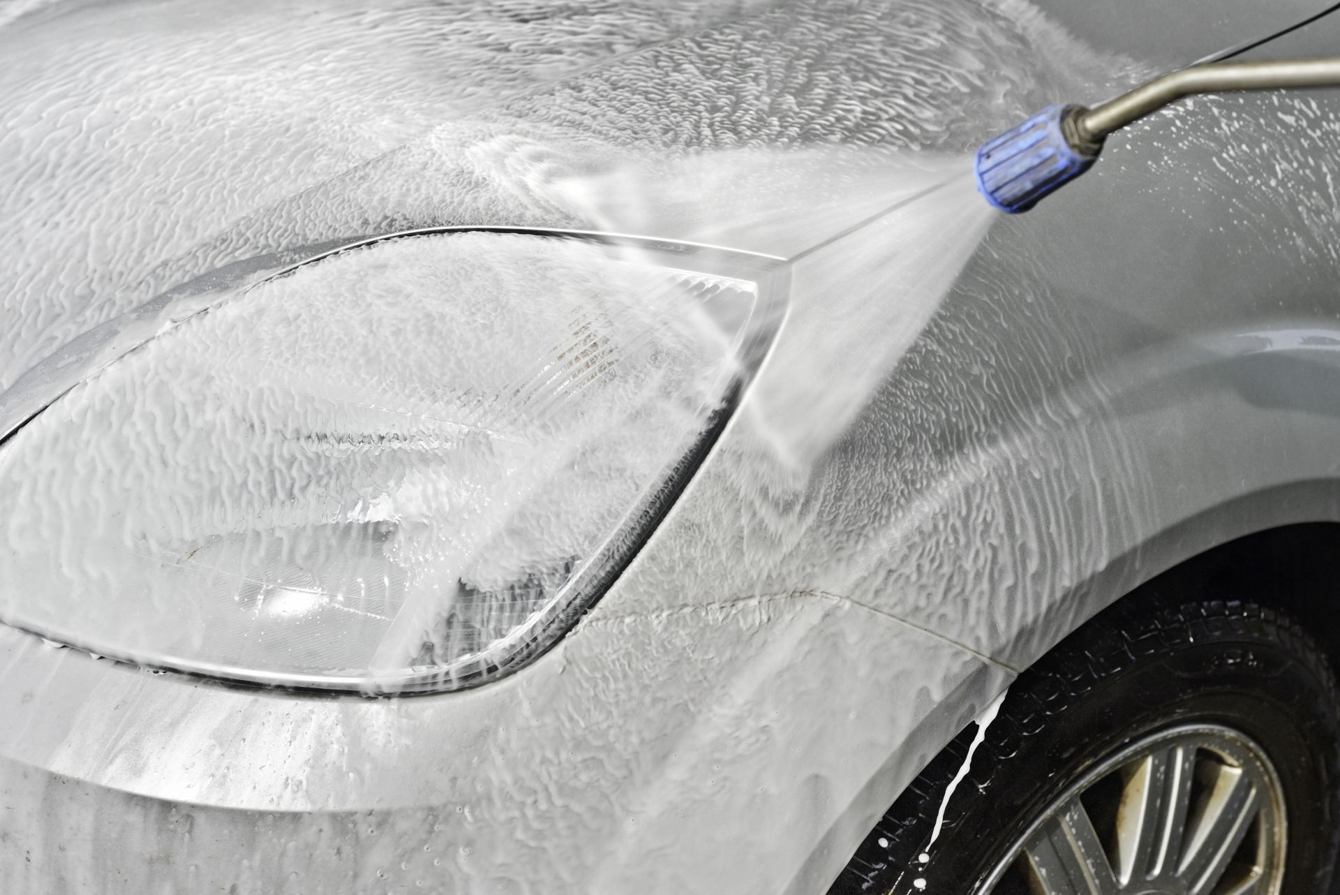 Car being washed with a pressure washer, spraying soapy water on the headlight and body.