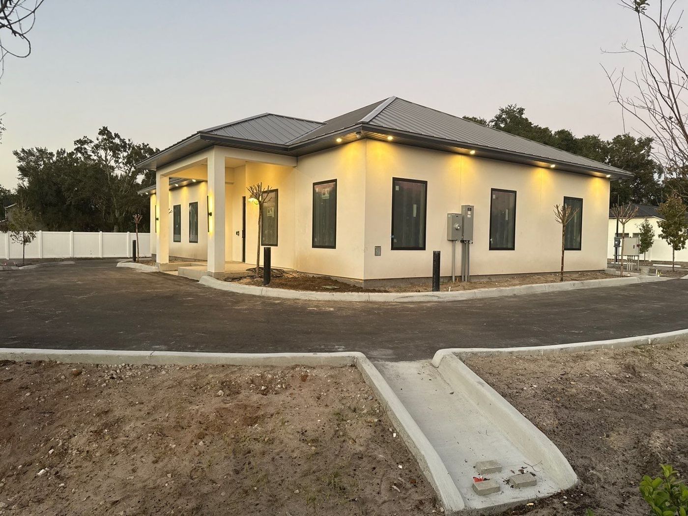 Small cream-colored house at dusk with lit windows and a gravel driveway in front.