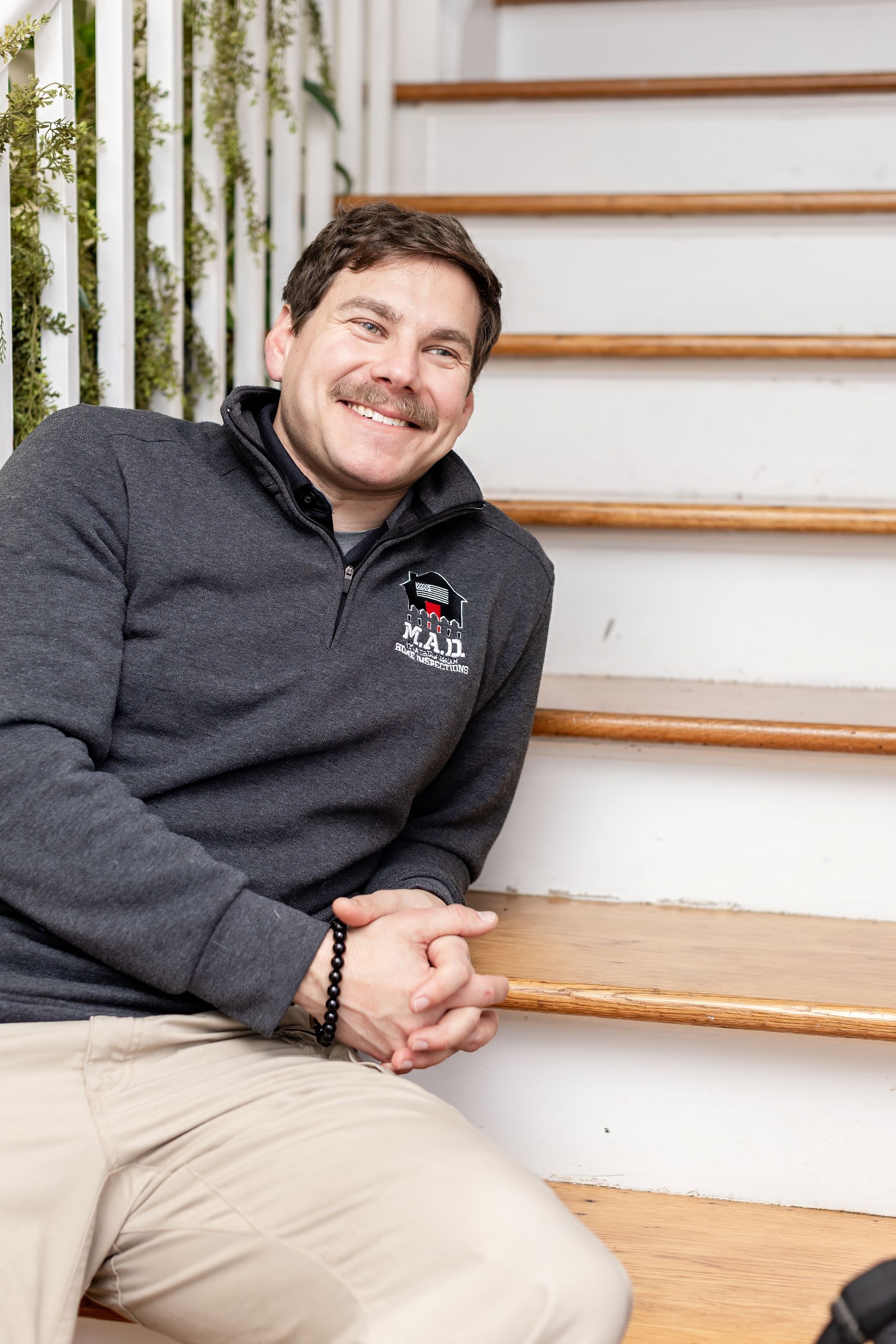 Smiling person in a dark sweater sitting on wooden steps beside a white staircase.