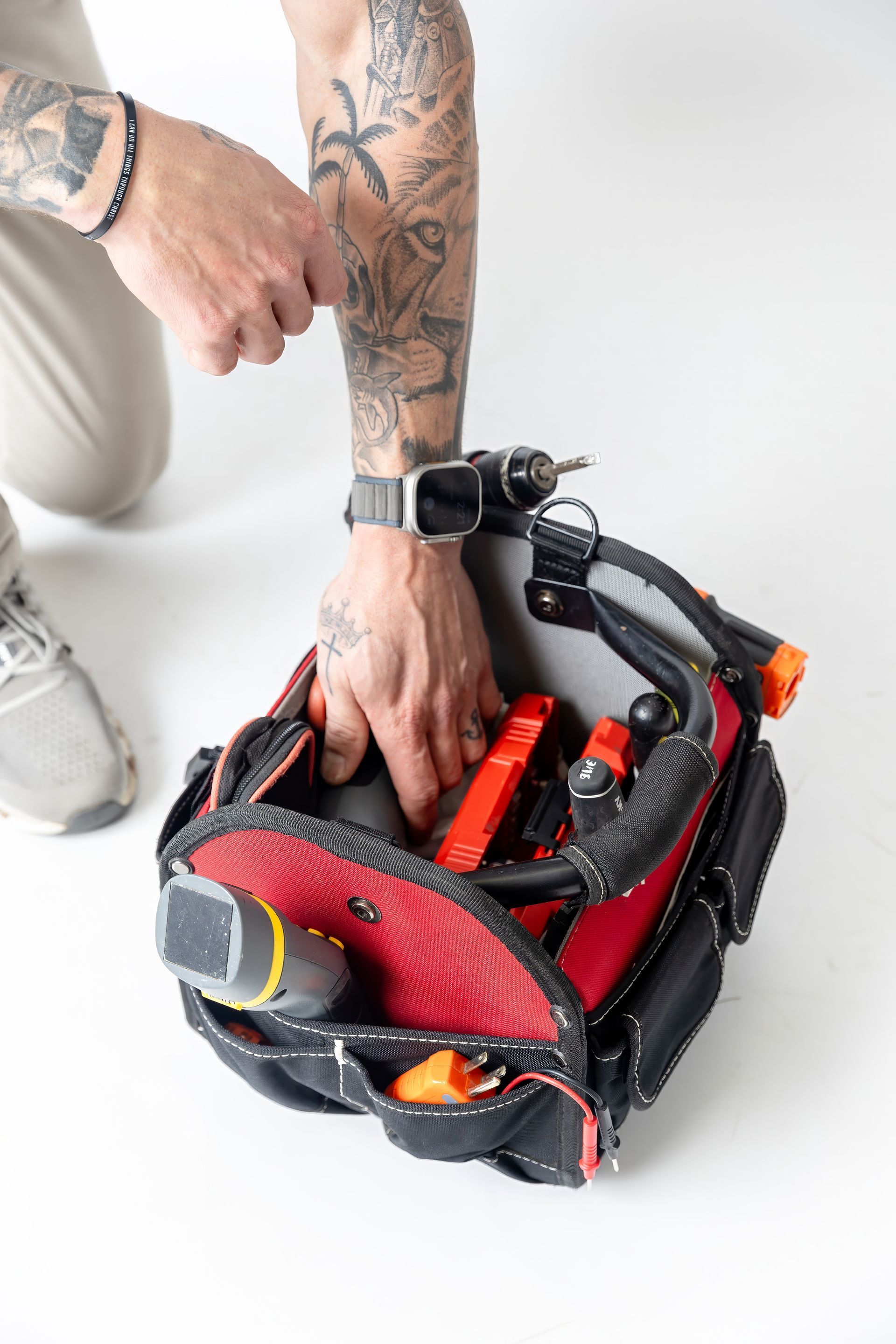 Tattooed arm packing tools into a black-and-red tool bag on the floor