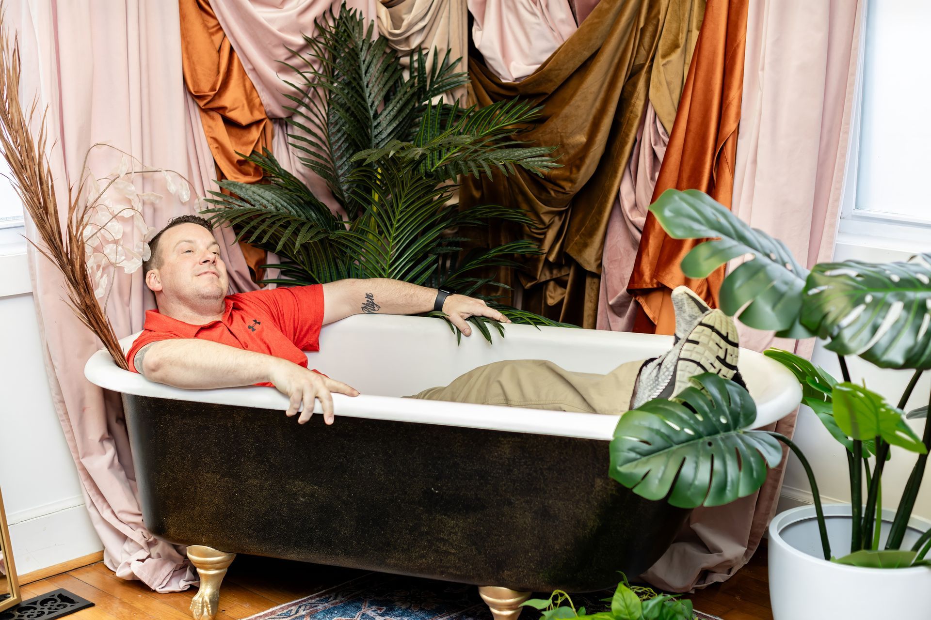 Person reclining in a black bathtub surrounded by lush houseplants and draped curtains.