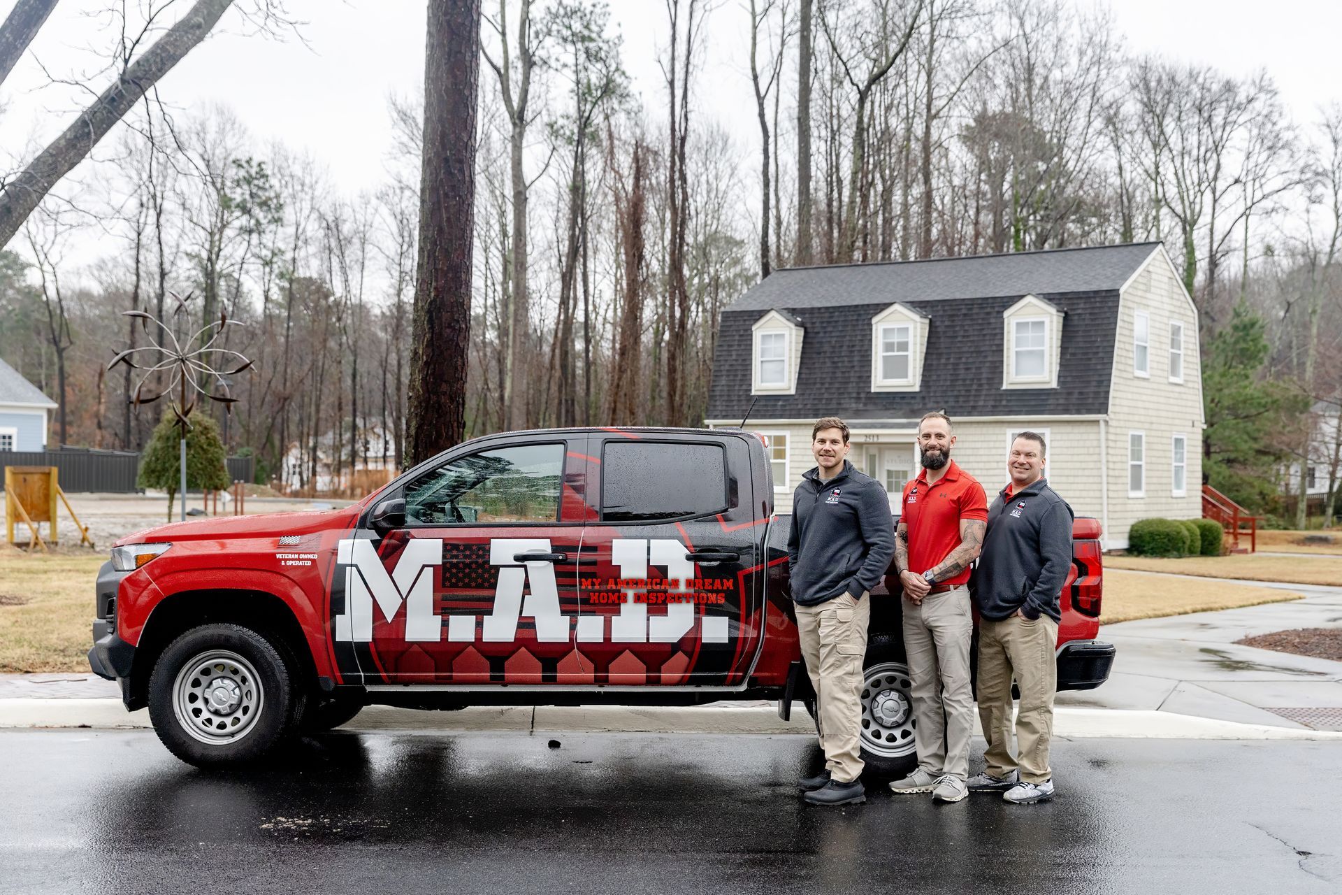 Three men posing beside a red-and-white M.J. Miller truck on a wet street, with a house and bare trees behind.