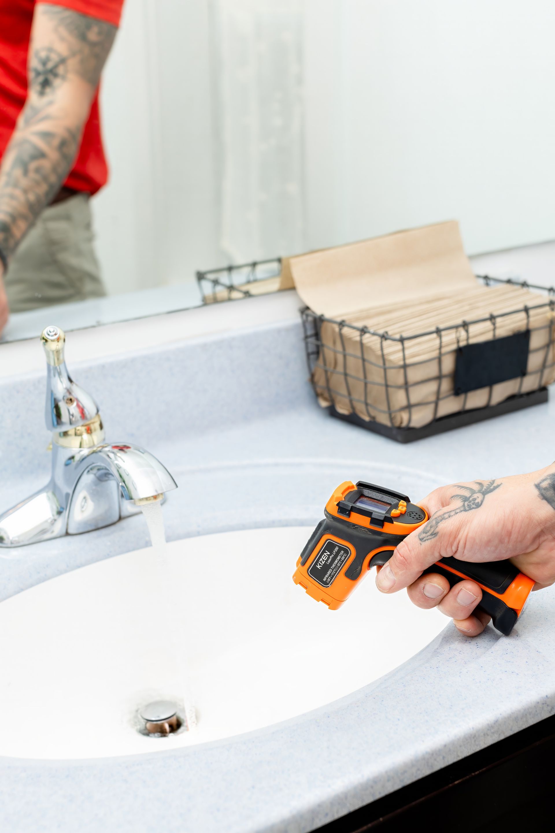 Hand holding an orange-and-black toy tool over a white bathroom sink beside a faucet and basket.