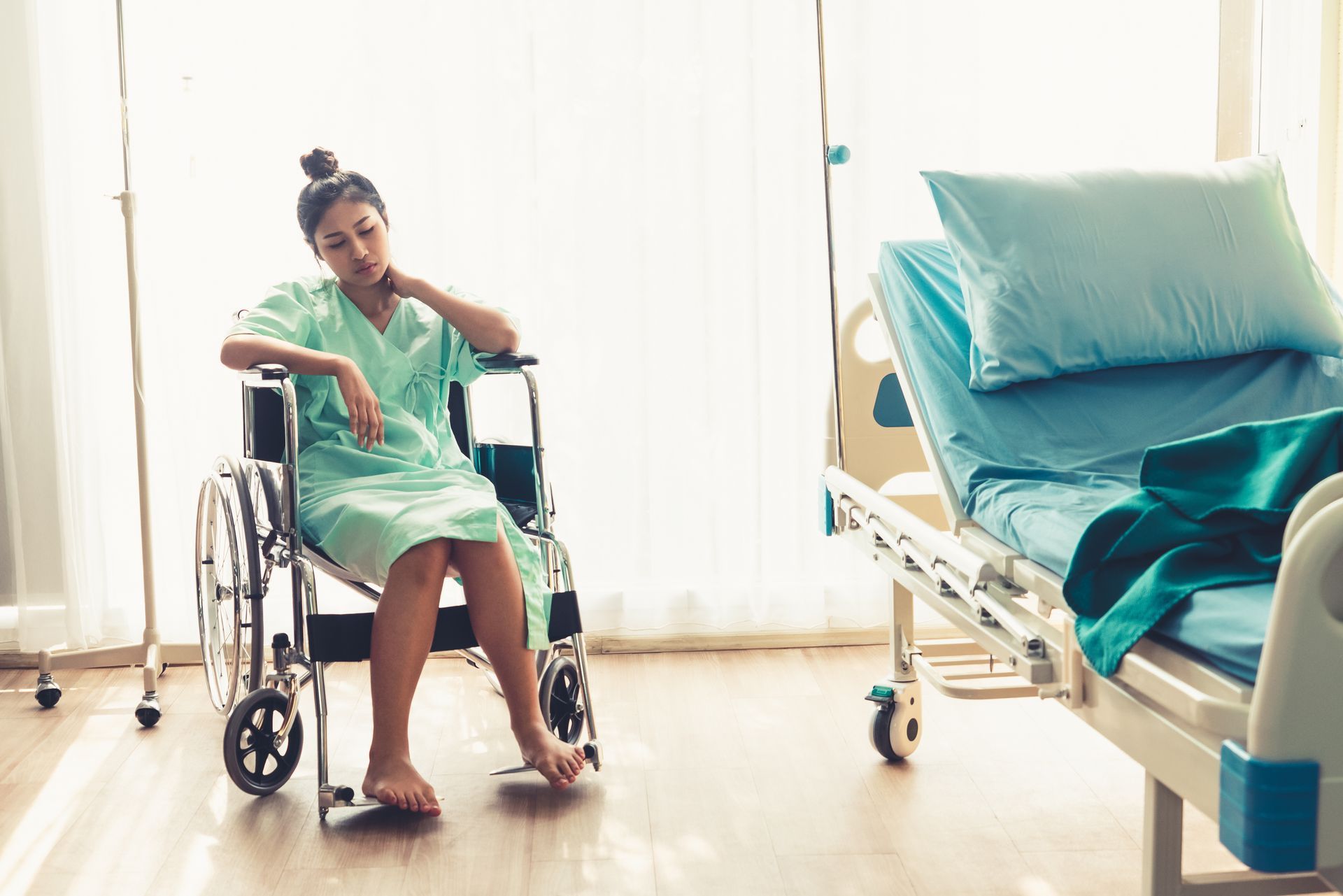 Woman in hospital gown sits in wheelchair, looking down. Hospital bed nearby, blue sheets.