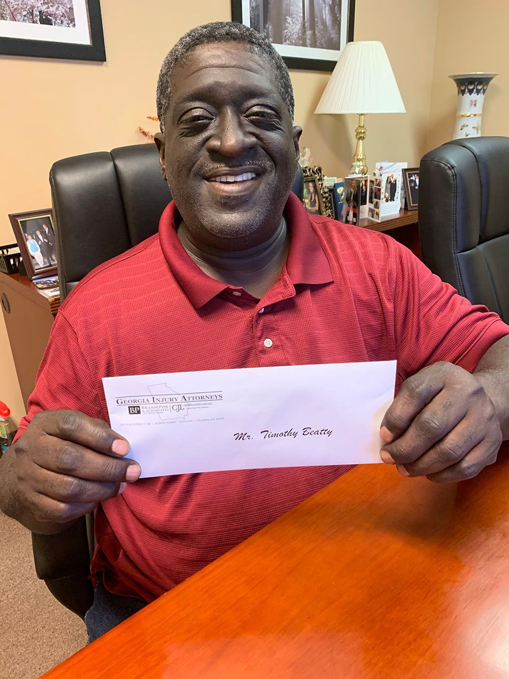 A man in a red shirt is sitting at a desk holding a check