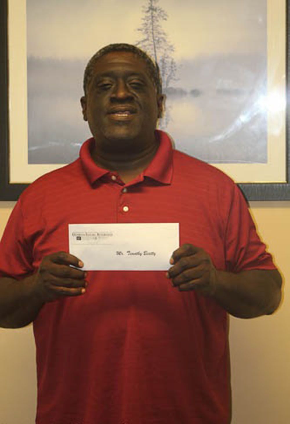 A man in a red shirt is holding a check in front of a picture