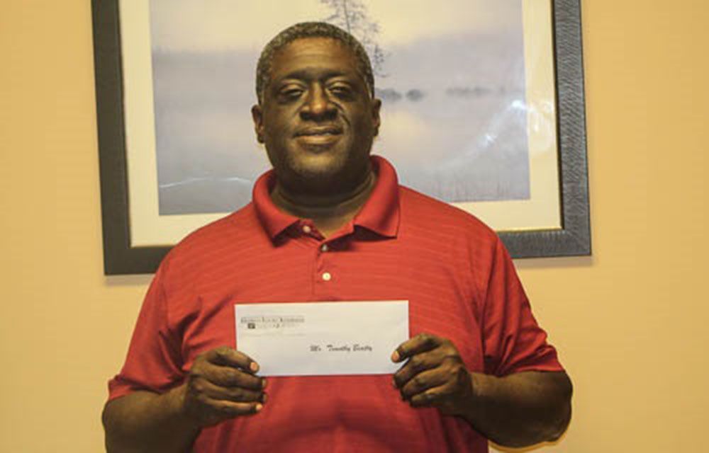 A happy man in a red shirt is holding a check in front of a picture