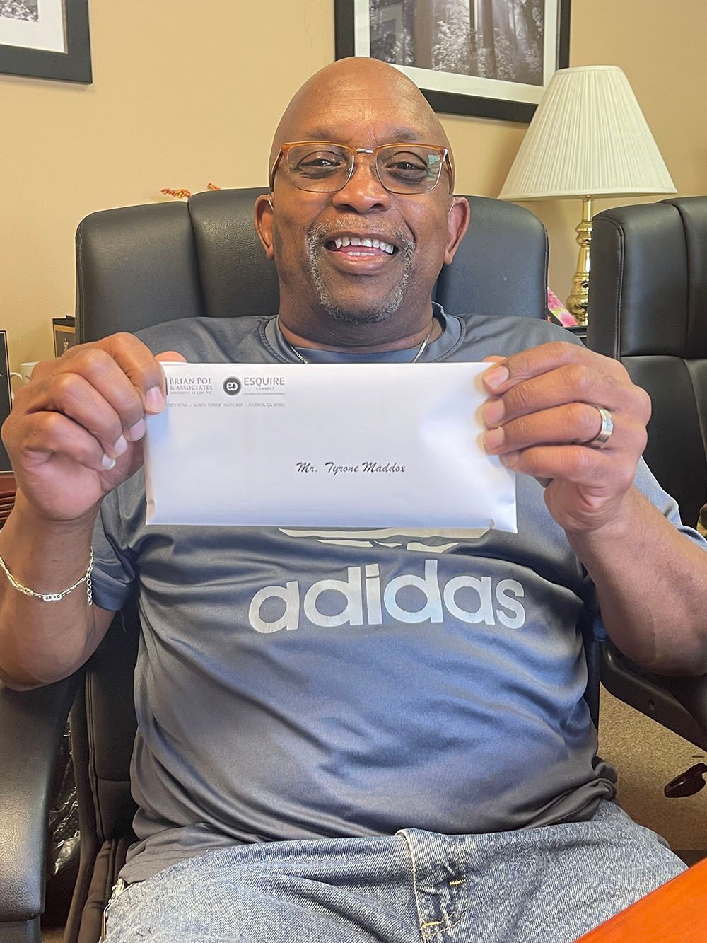A man in an Adidas shirt is sitting in a chair holding a check