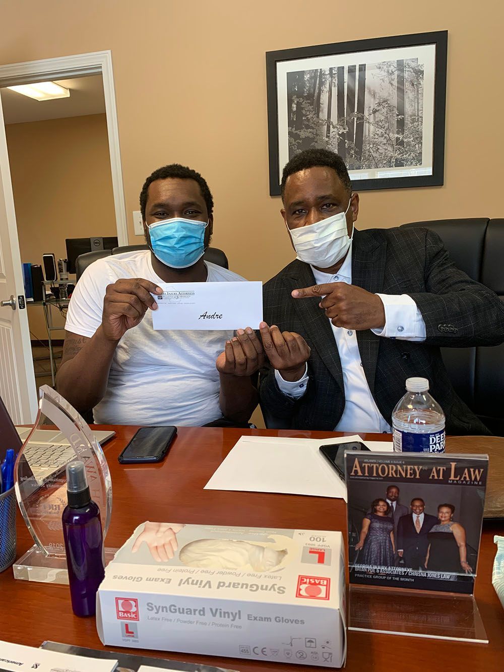 Brian D. Poe, Esq. and a client wearing face masks are sitting at a desk holding a check