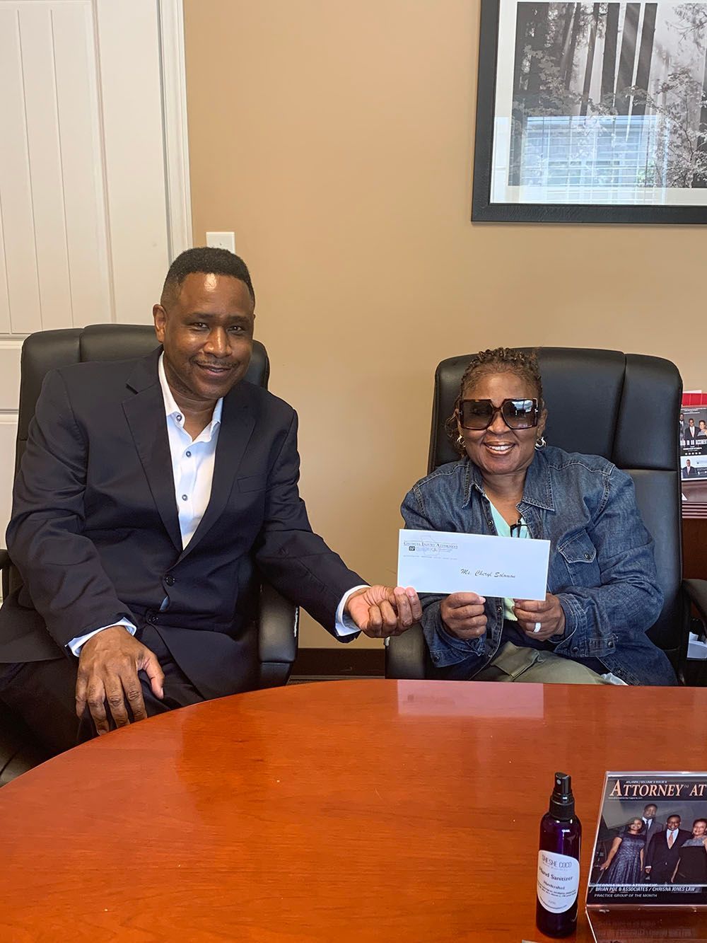 Brian D. Poe, Esq. and a woman in a denim jacket while wearing an eyeglasses are sitting at a table holding a check