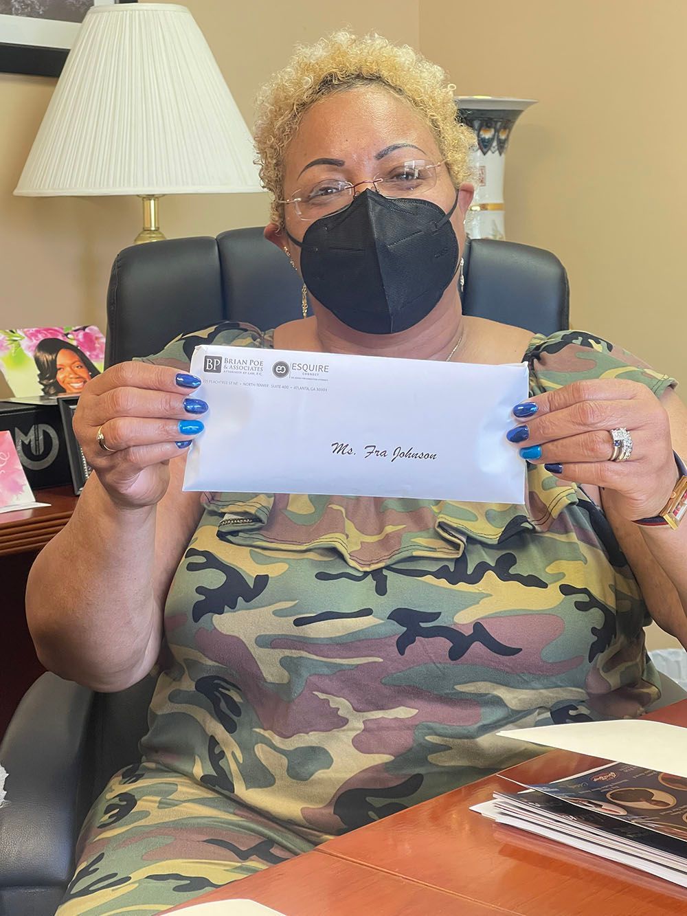 A happy woman wearing a mask is sitting at a desk holding a check