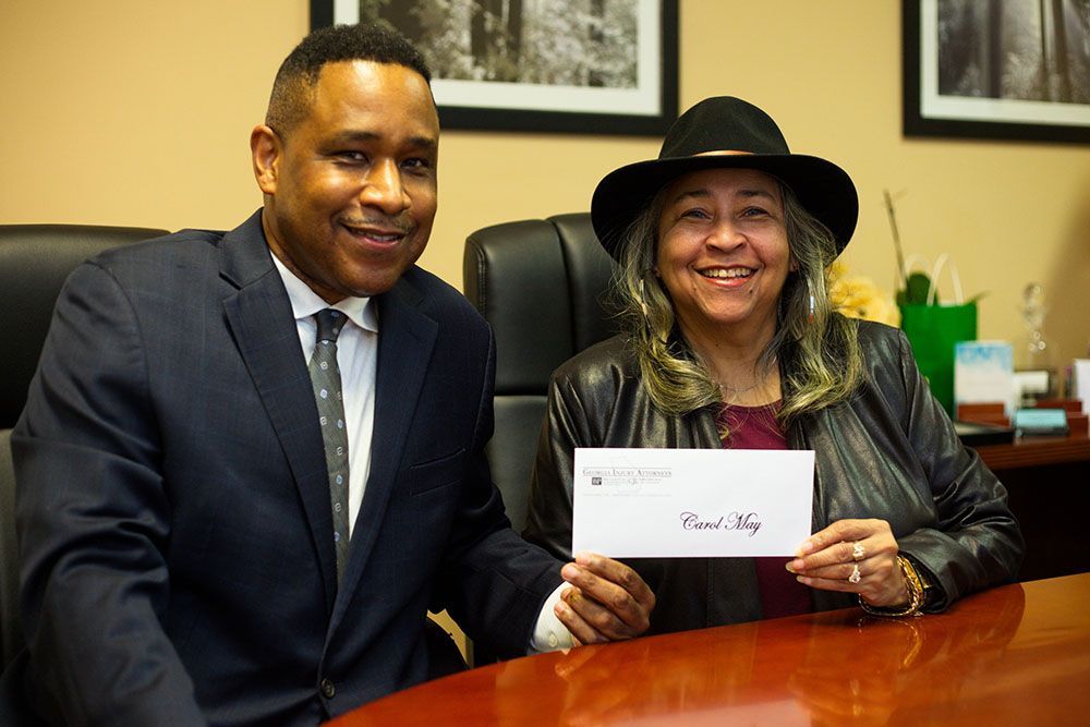 Brian D. Poe, Esq. and a happy woman wearing a hat are sitting at a table holding a check