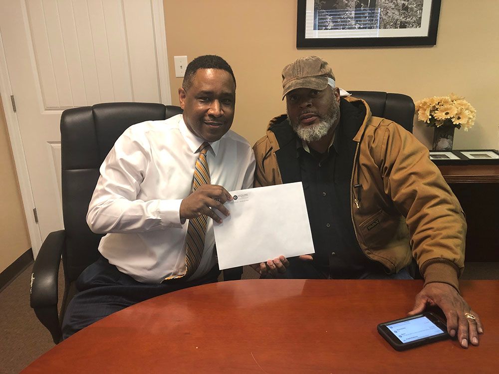 Brian D. Poe, Esq. and a client wearing a hat and jacket are sitting at a table holding a check