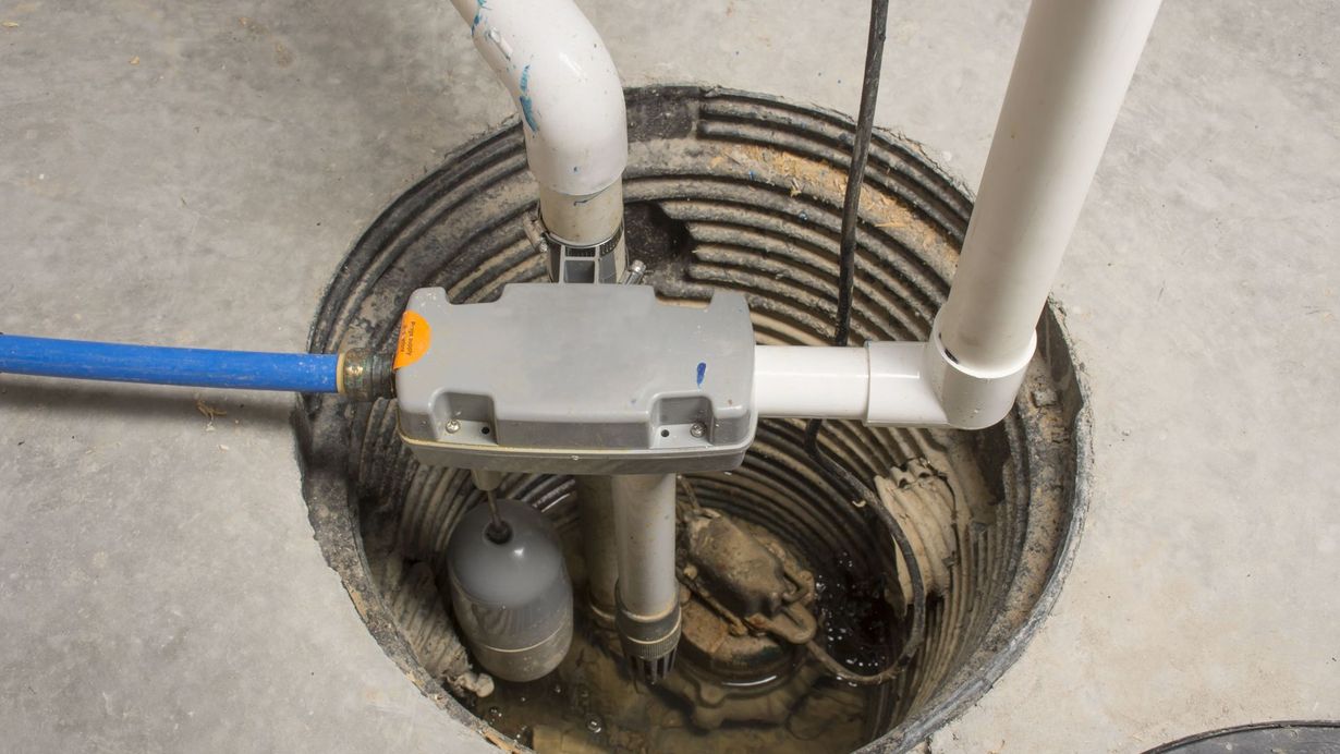 Open sump pit with pipes, wiring, and a gray control box in a concrete floor