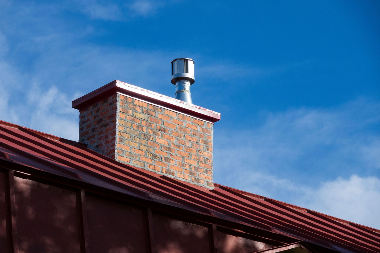 Rooftop chimney with metal vent against a blue sky above red roof tiles