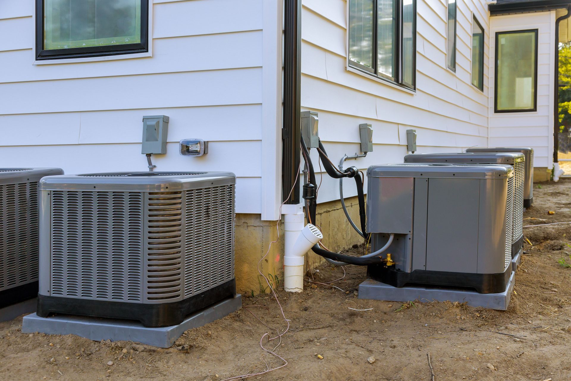 Two outdoor HVAC units beside a white house, with dirt ground and siding visible.
