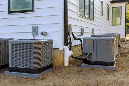 Two outdoor air conditioning units beside a house, with white siding and a dirt yard.