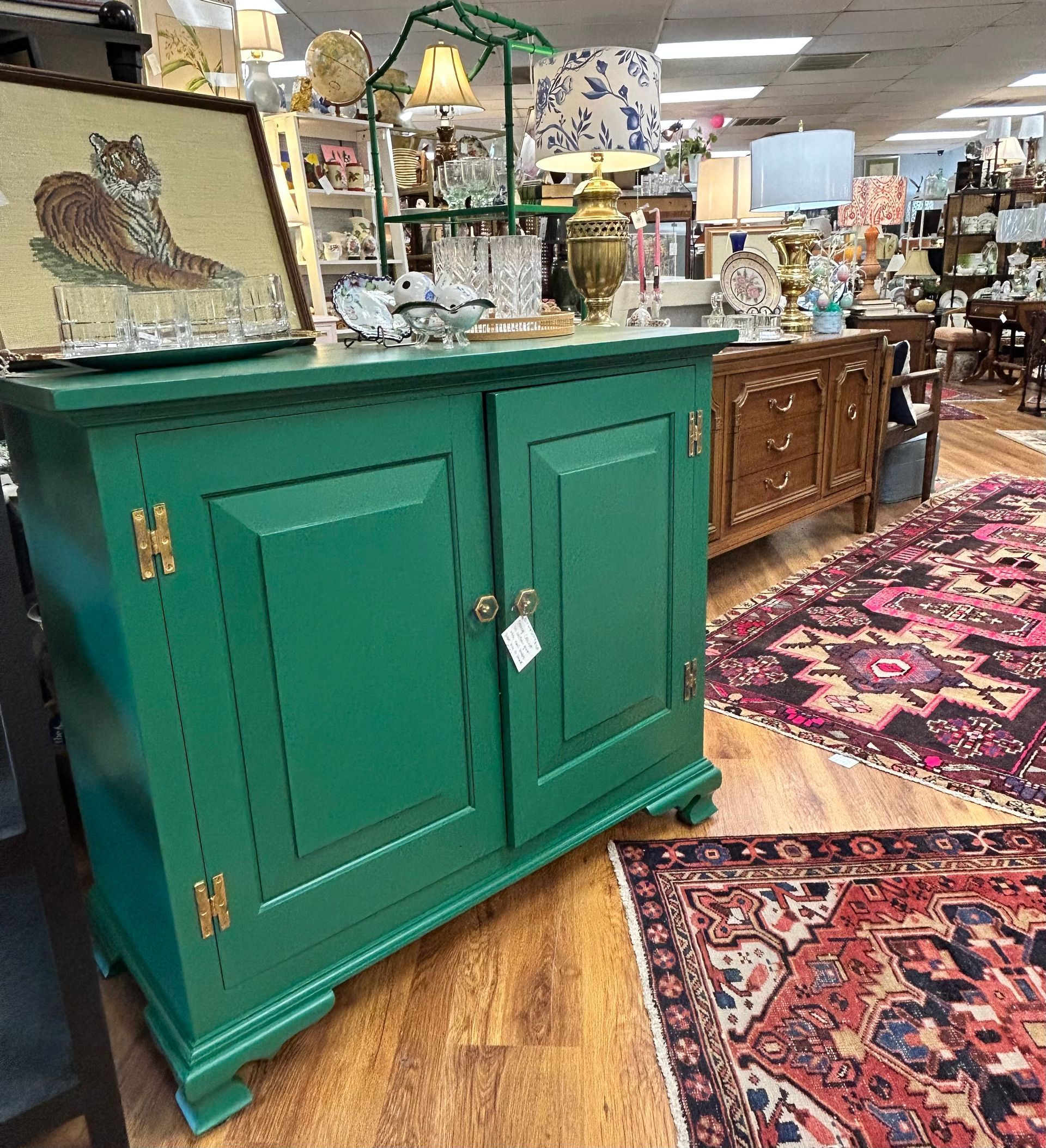 A vibrant green wooden cabinet with brass hinges stands in a store, surrounded by antique lamps and patterned rugs.