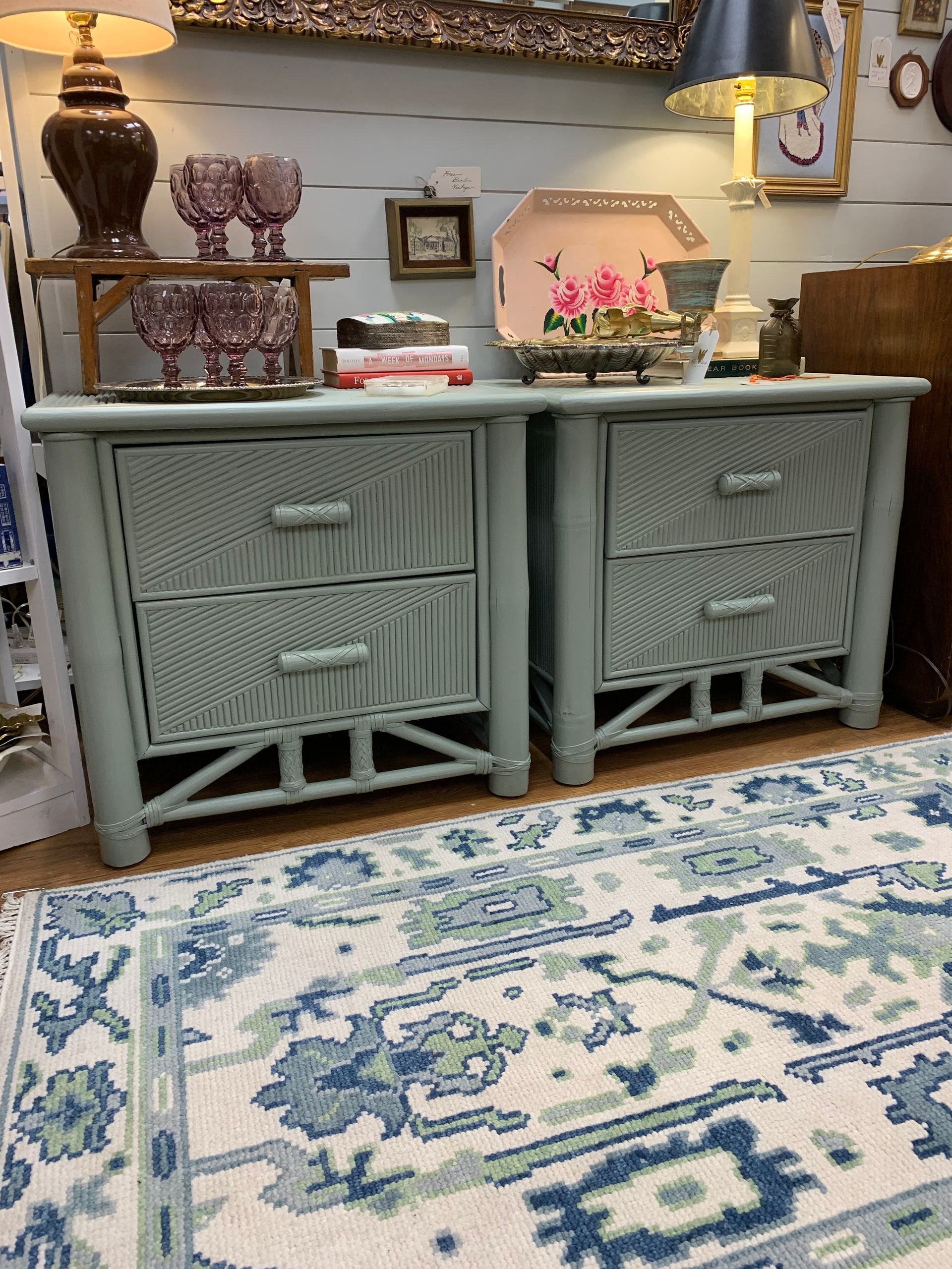 Two pale green, wicker-textured nightstands with drawers sit on a patterned blue and white area rug in a room.