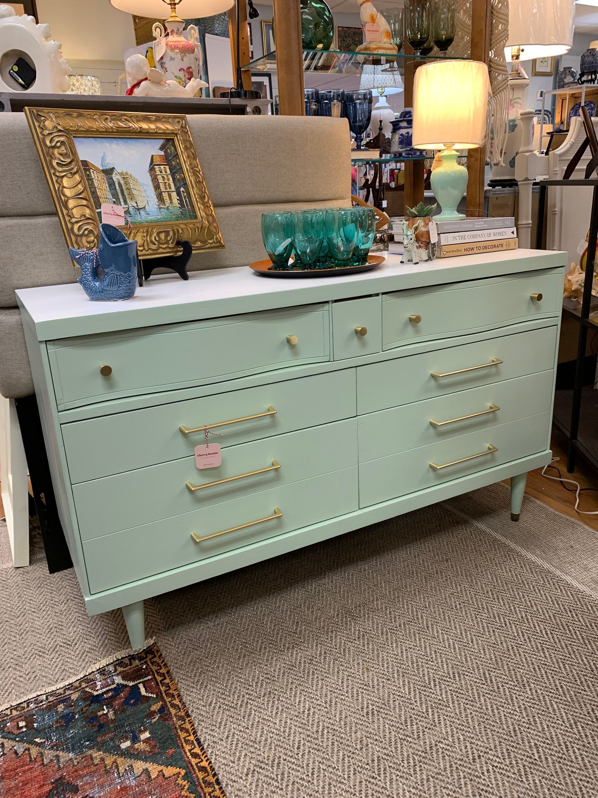 A mint green mid-century dresser with gold hardware and detailed trim sits on a rug in a furniture store.