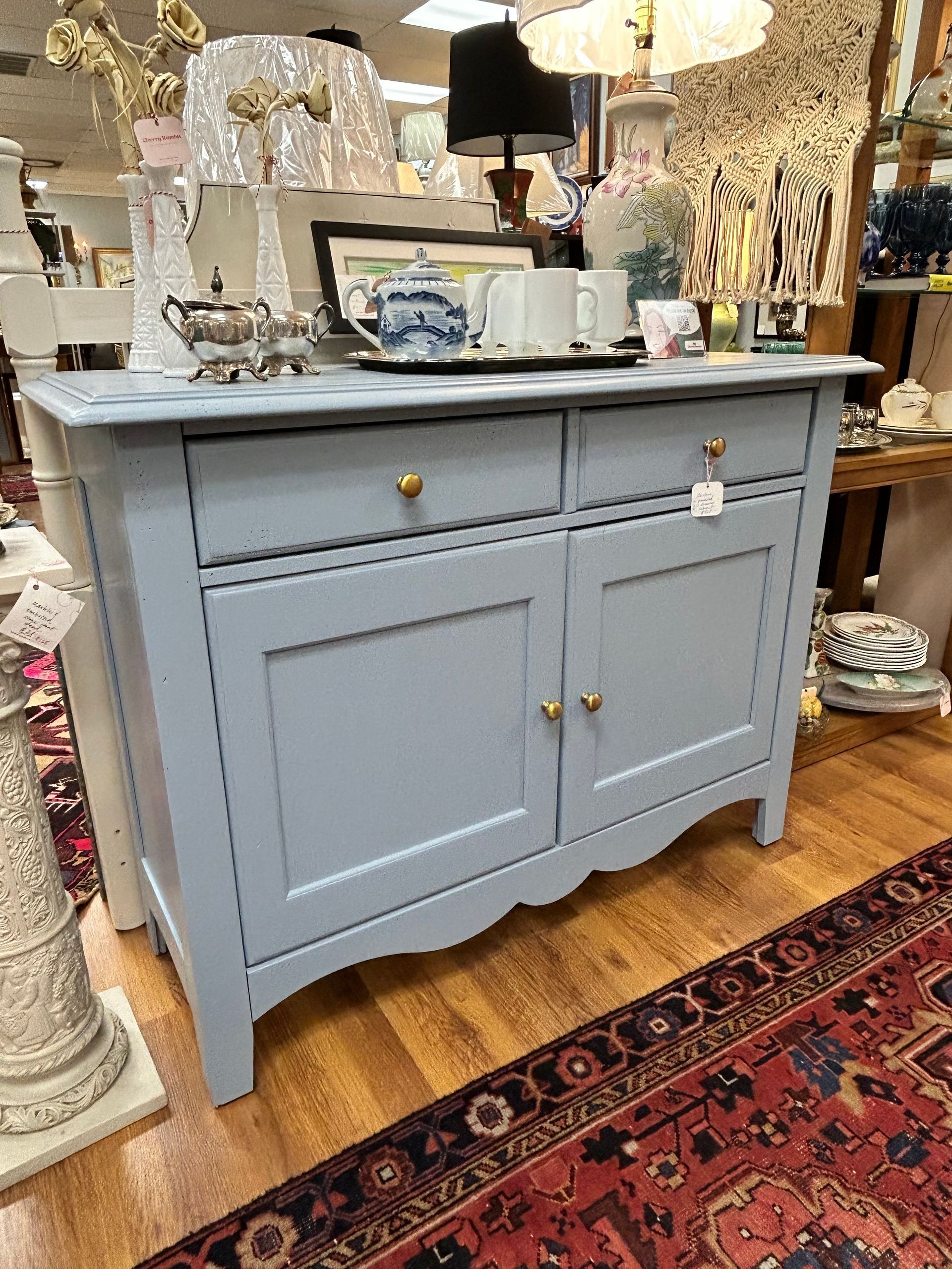 A light blue, two-drawer, two-door wooden sideboard with brass knobs sitting on a wooden floor in a shop.