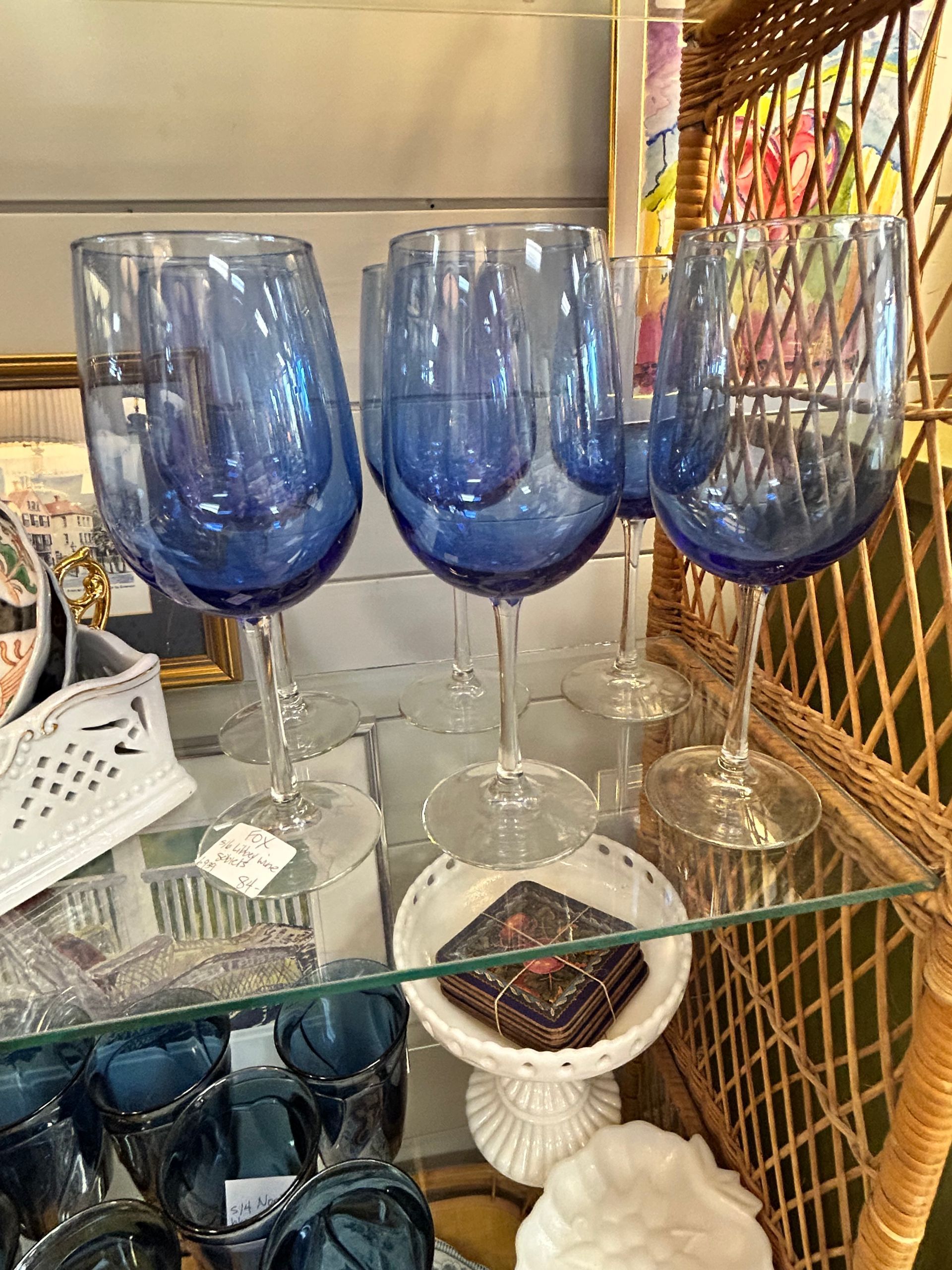 Three cobalt blue wine glasses are displayed on a glass shelf in a retail setting, with some blurred glassware below.