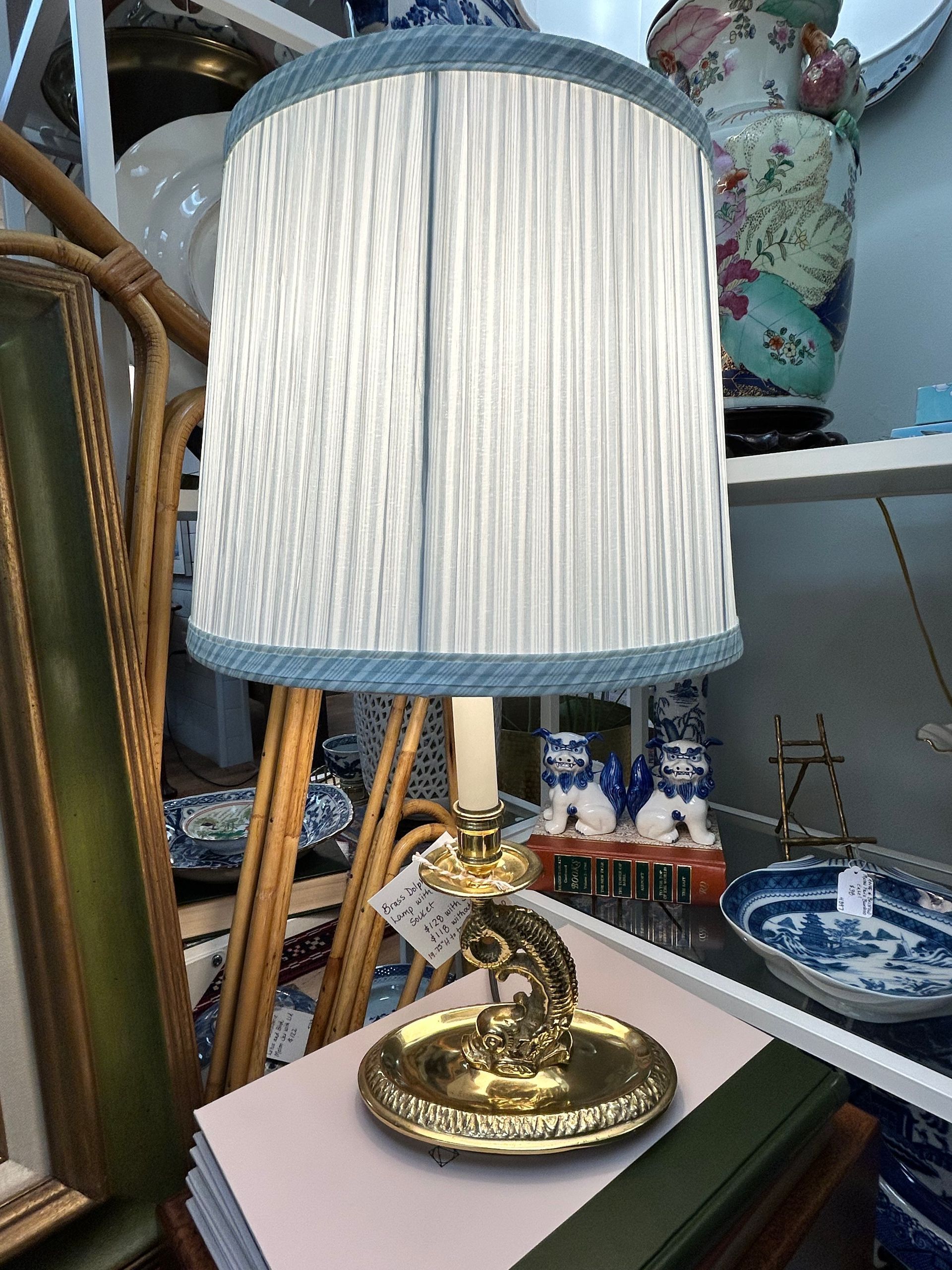 A brass candlestick lamp with a pleated, light blue-trimmed lampshade sits on a book in a shop with various collectibles.