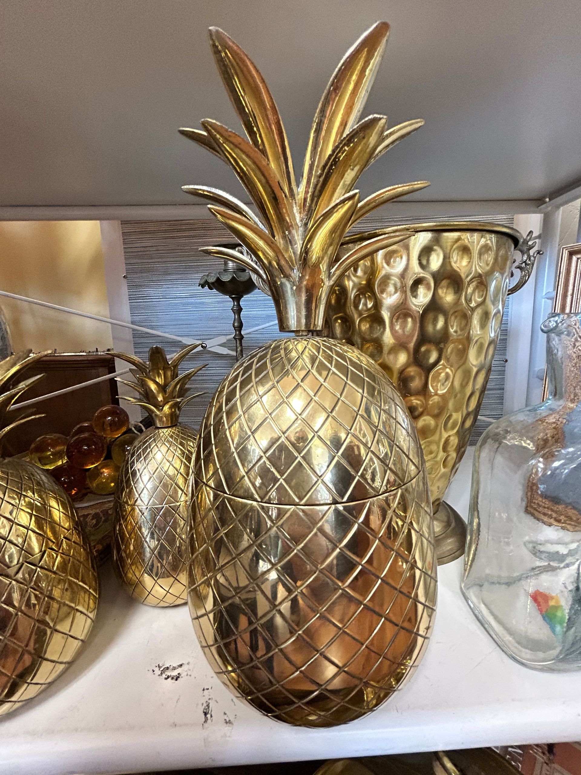 Three metallic, gold-colored pineapple-shaped containers or sculptures sit on a white shelf.