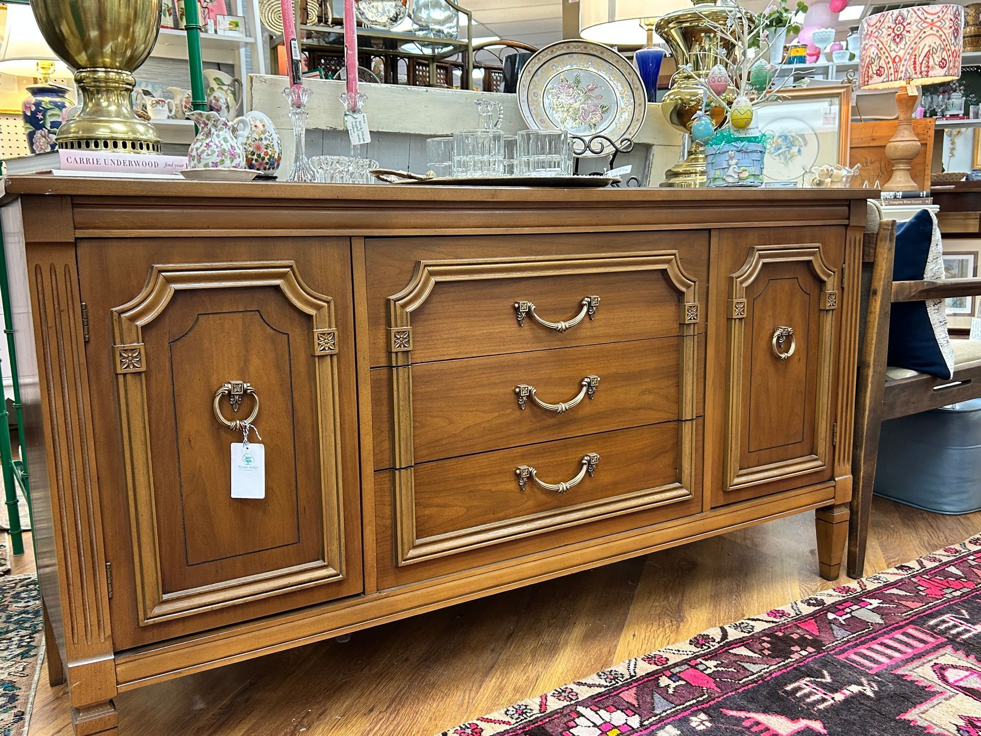 A wooden sideboard with two side cabinet doors and three central drawers featuring ornate gold-toned pull hardware.