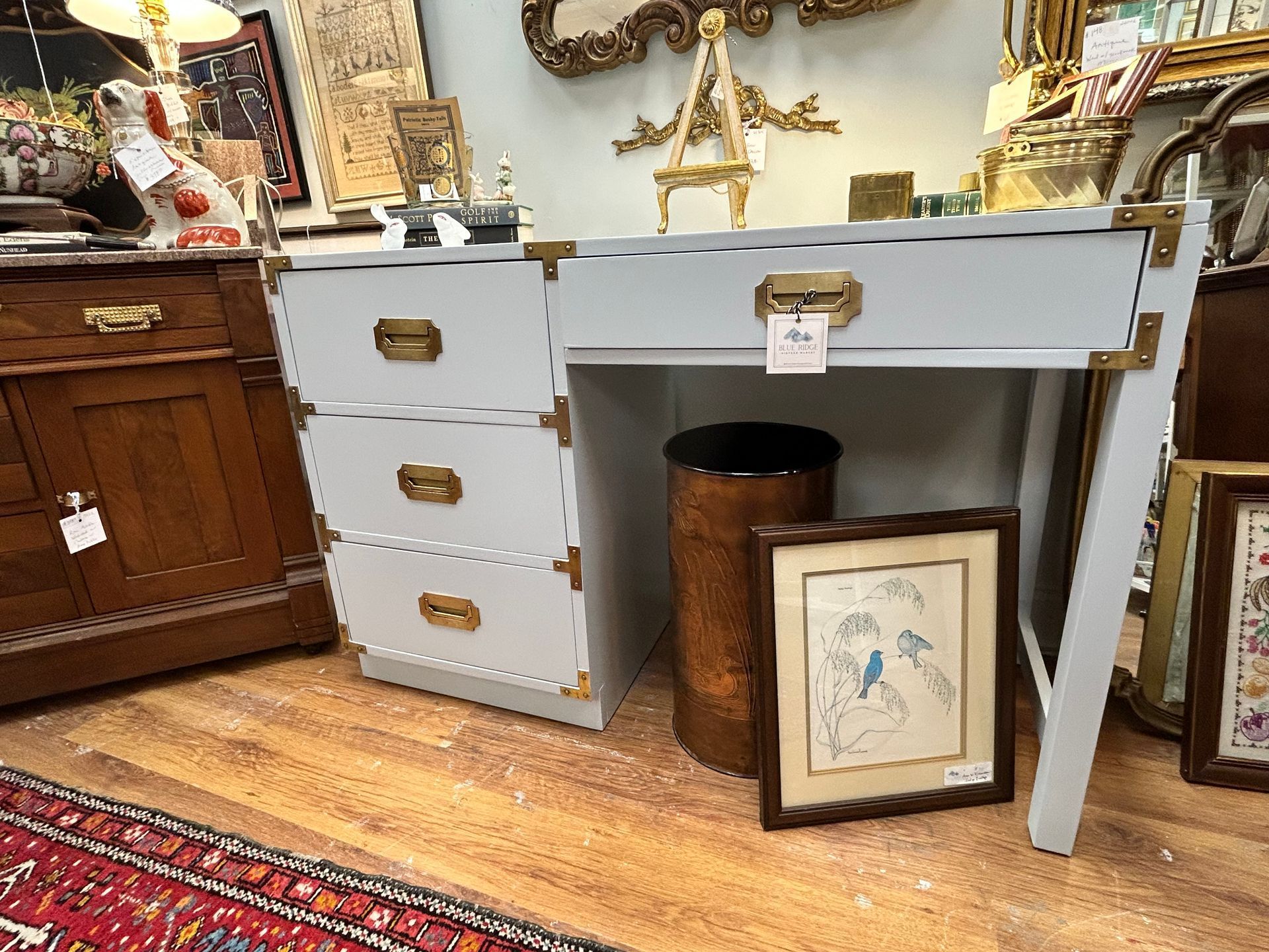 A light blue campaign-style desk with brass hardware stands on a wood floor near a framed print and a metal trash bin.