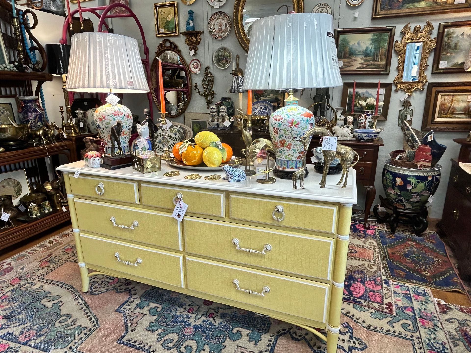 A yellow six-drawer dresser with white accents and two lamps, displayed in an antique shop filled with wall decorations.