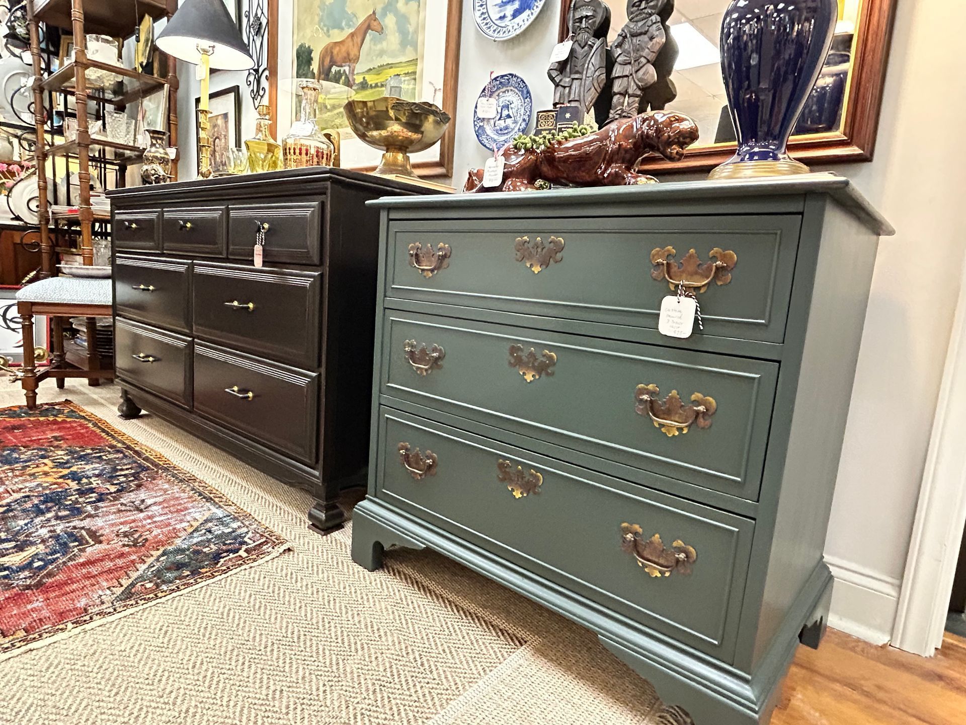 Two dressers are on display in a store, one painted dark green with gold hardware, and a taller black one behind it.
