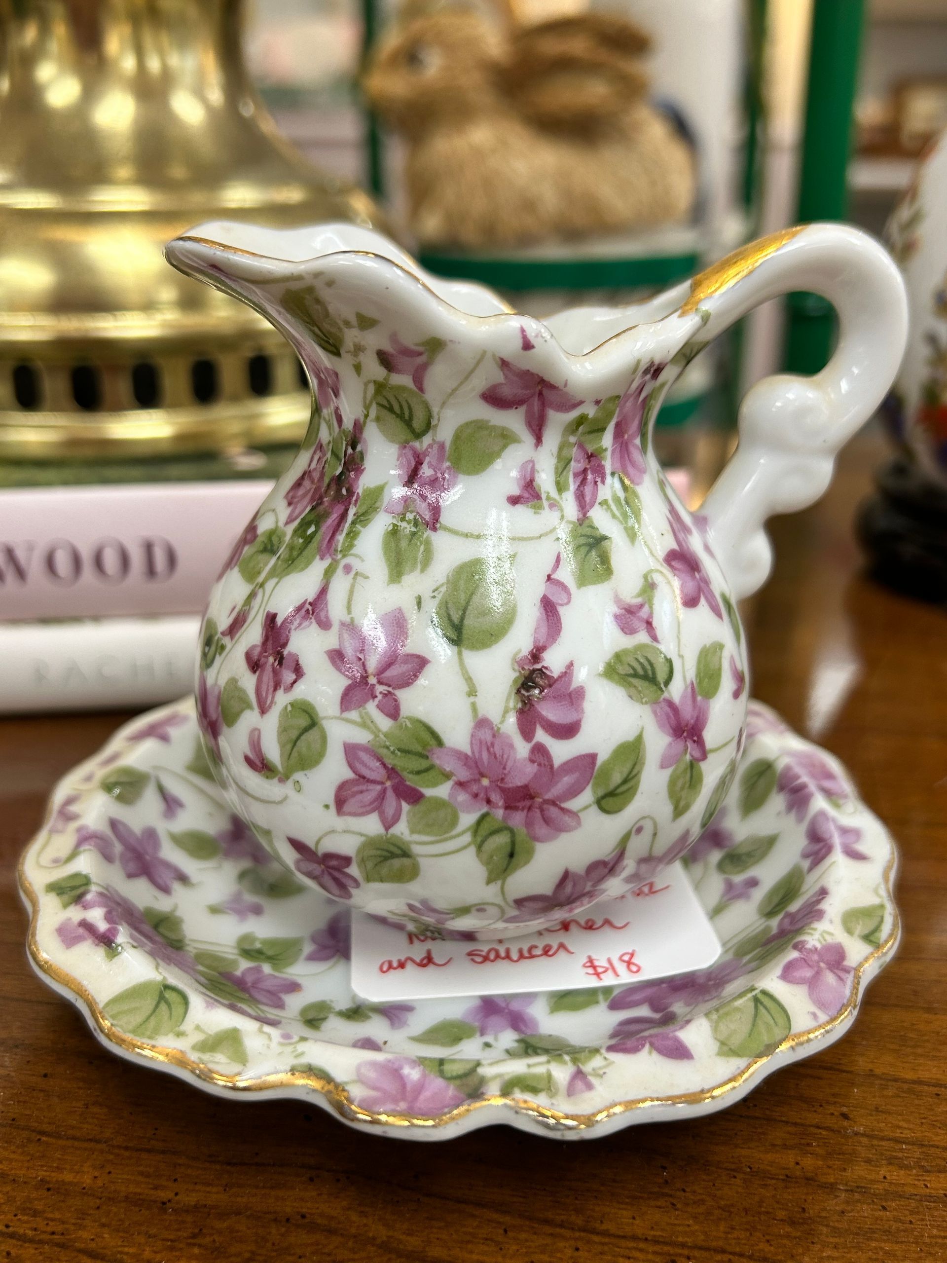 A vintage porcelain creamer and saucer set decorated with purple flowers, green leaves, and gold trim.