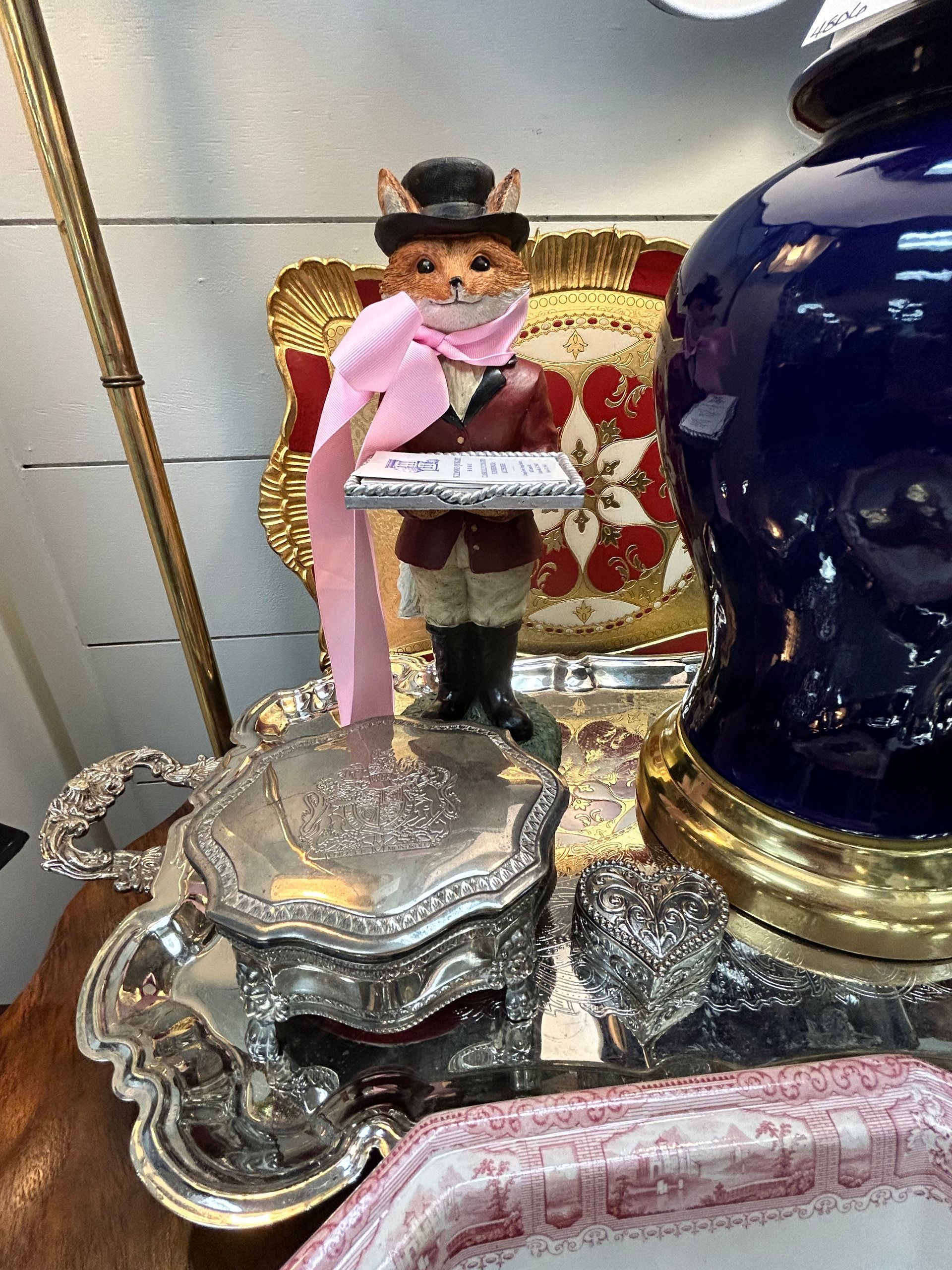 A whimsical fox figurine in a top hat and scarf stands on a decorative silver tray next to a large dark blue lamp.