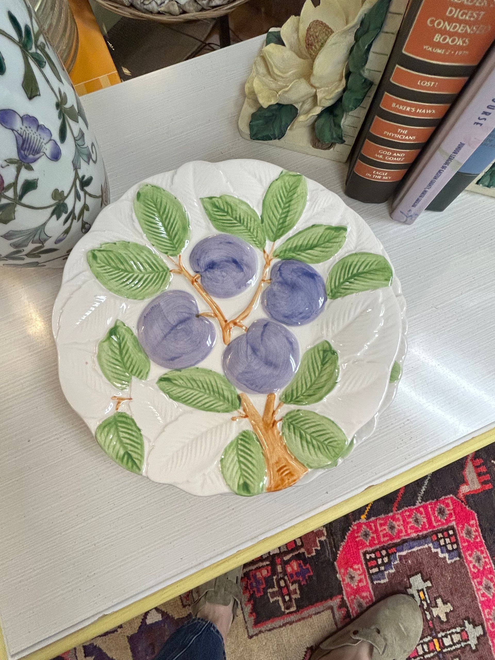 A white ceramic plate embossed with a branch of green leaves and four purple plums, sitting on a surface.