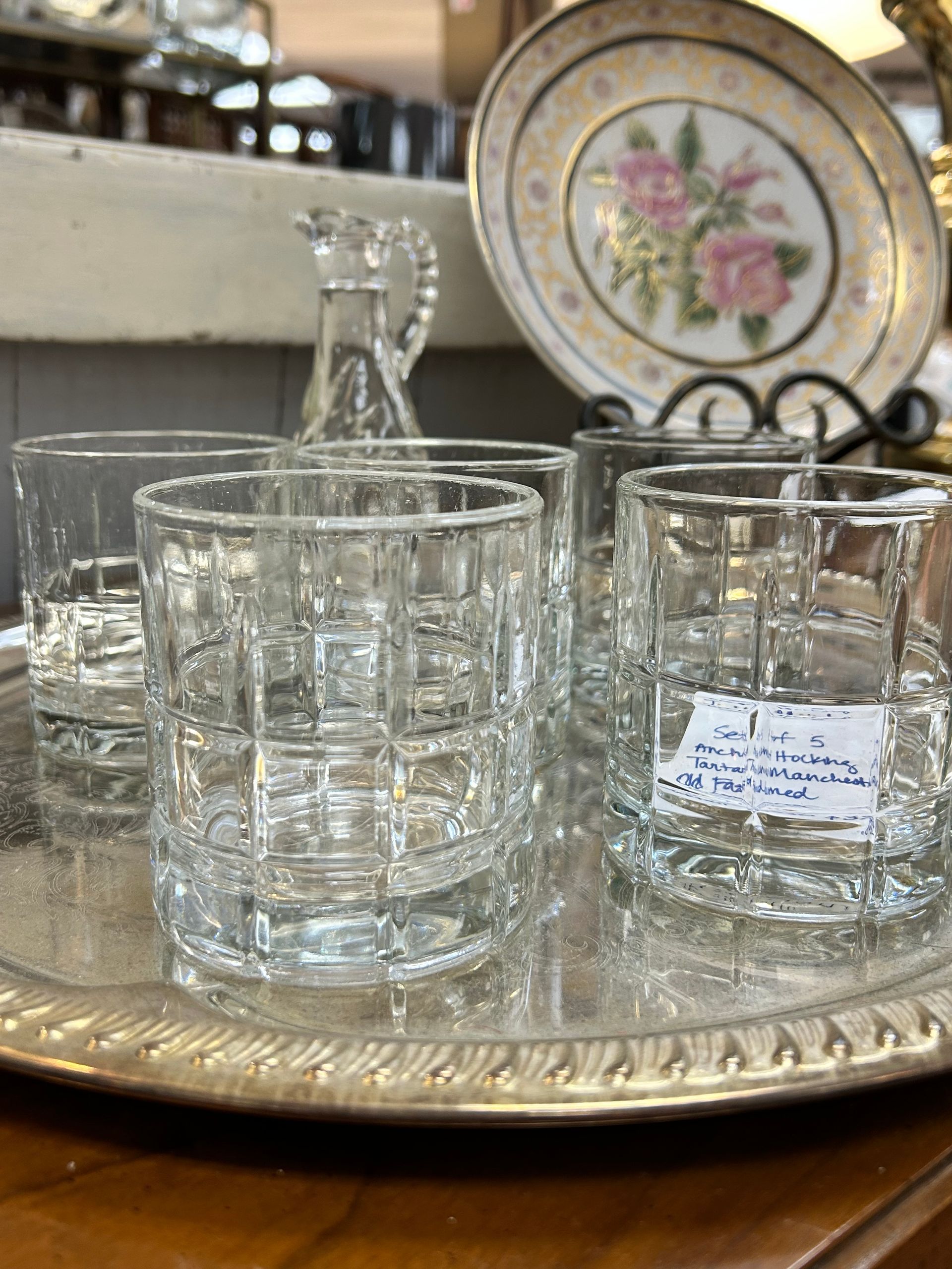A silver serving tray holds four clear, grid-patterned drinking glasses and a small glass carafe, with a floral plate behind.
