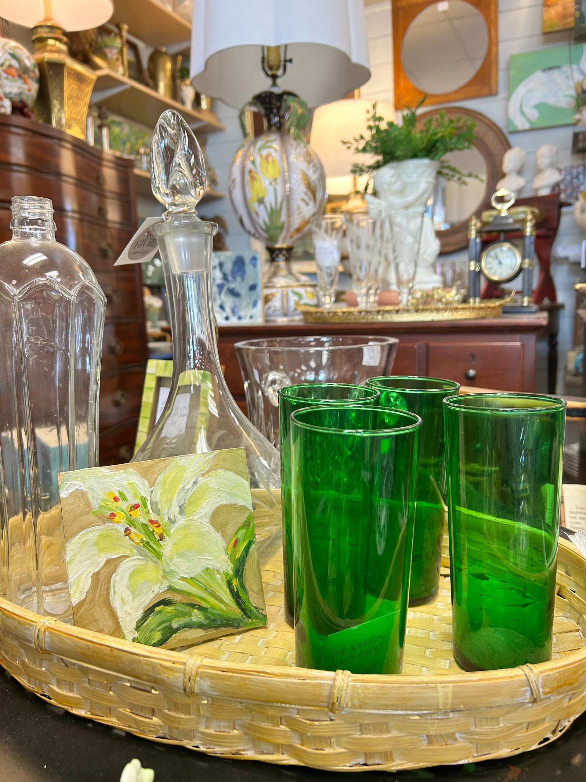 A shallow, woven basket holds a clear glass decanter, a decorative lily plaque, and three vibrant green drinking glasses.