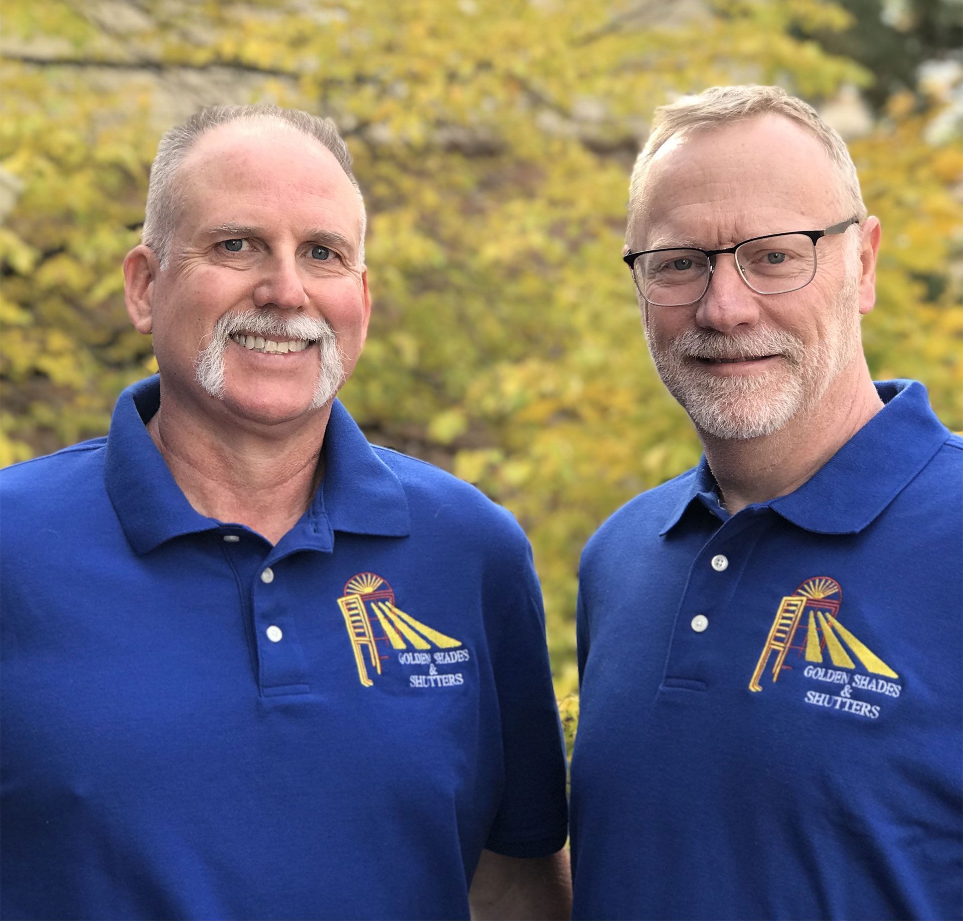 Two men in blue shirts pose outdoors, smiling. Their shirts have a logo. Yellow leaves in background.