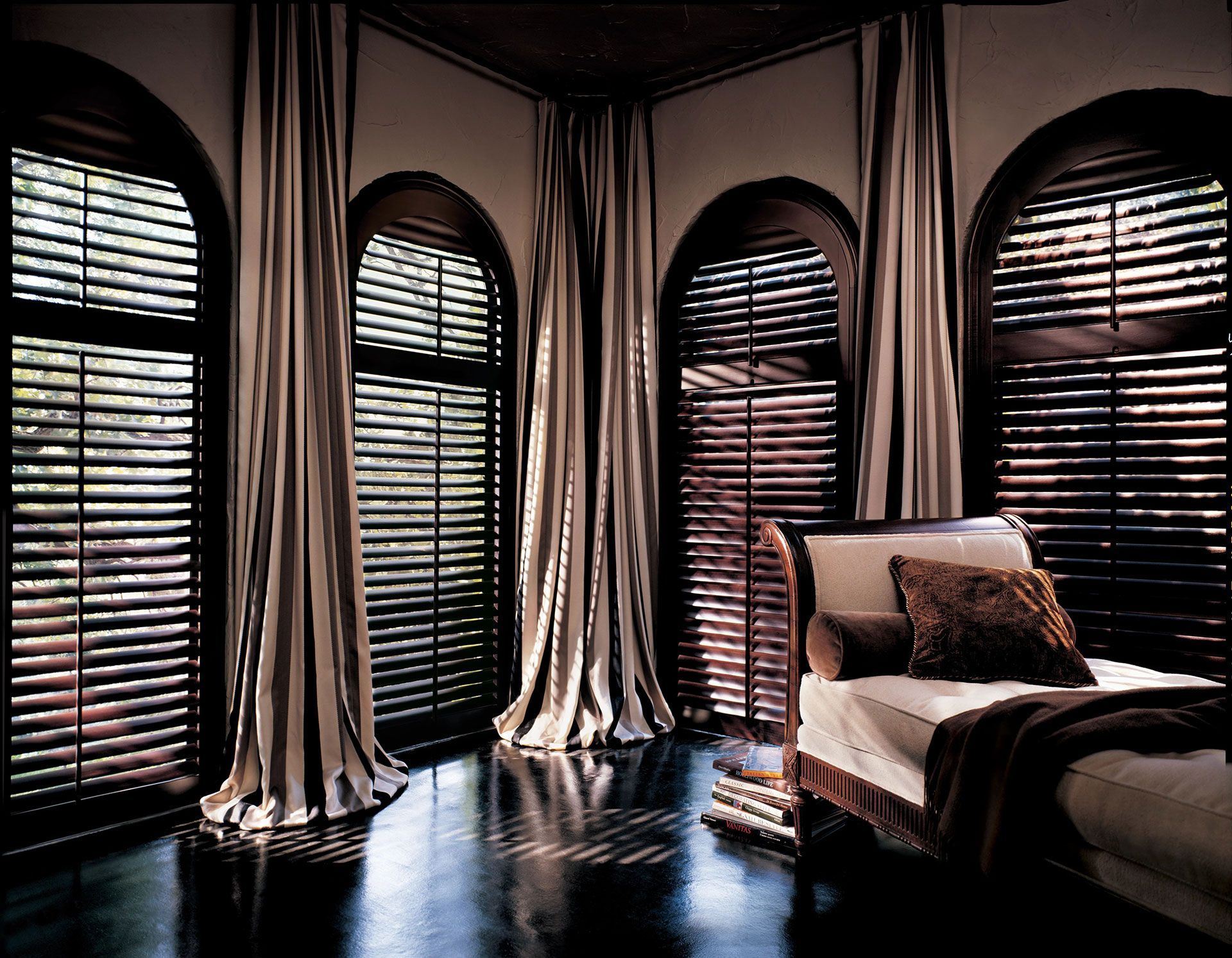 Bedroom with arched windows, dark shutters, beige curtains, and a chaise lounge.