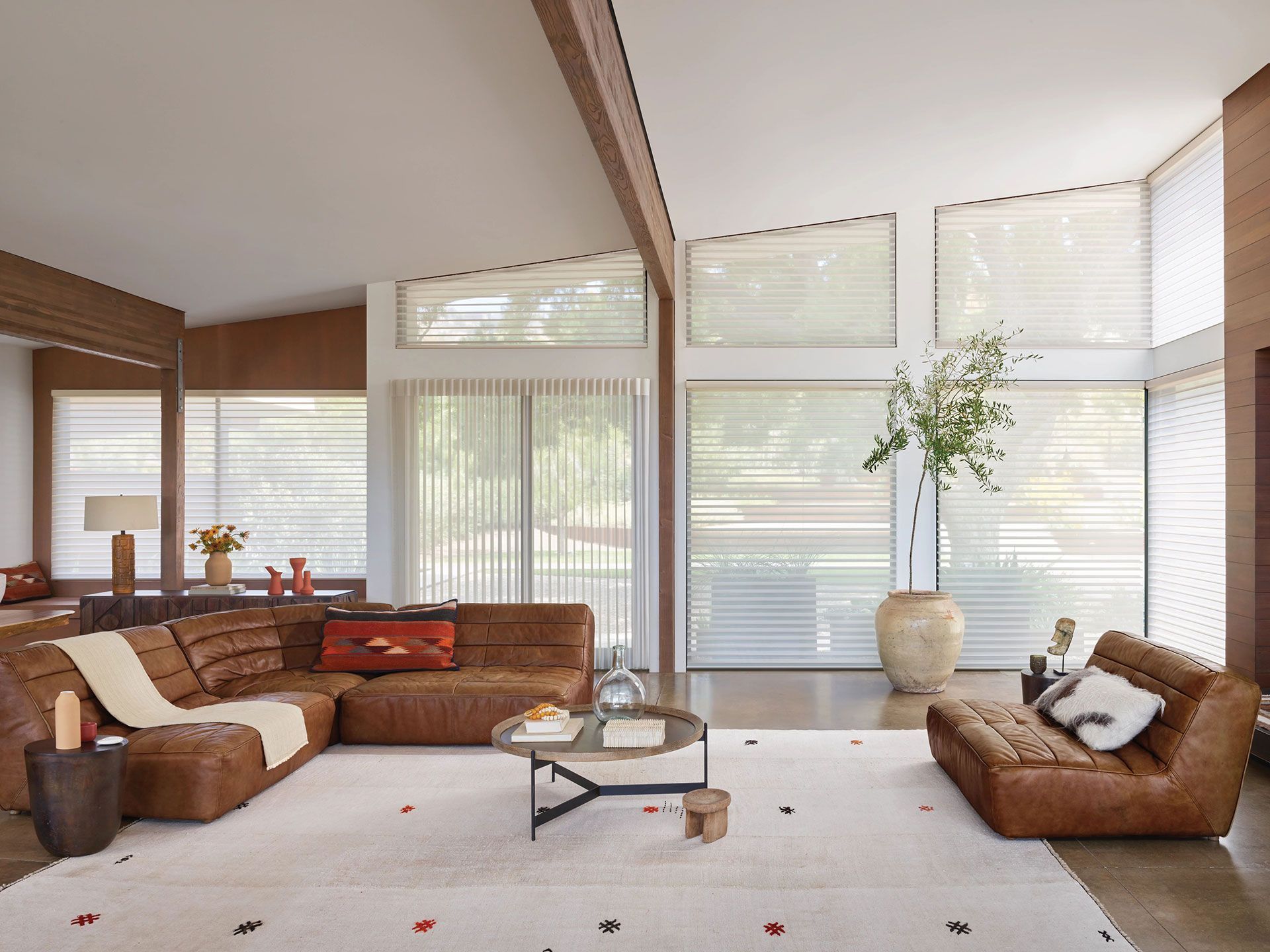 A modern living room with brown leather furniture, large windows, and a neutral rug.