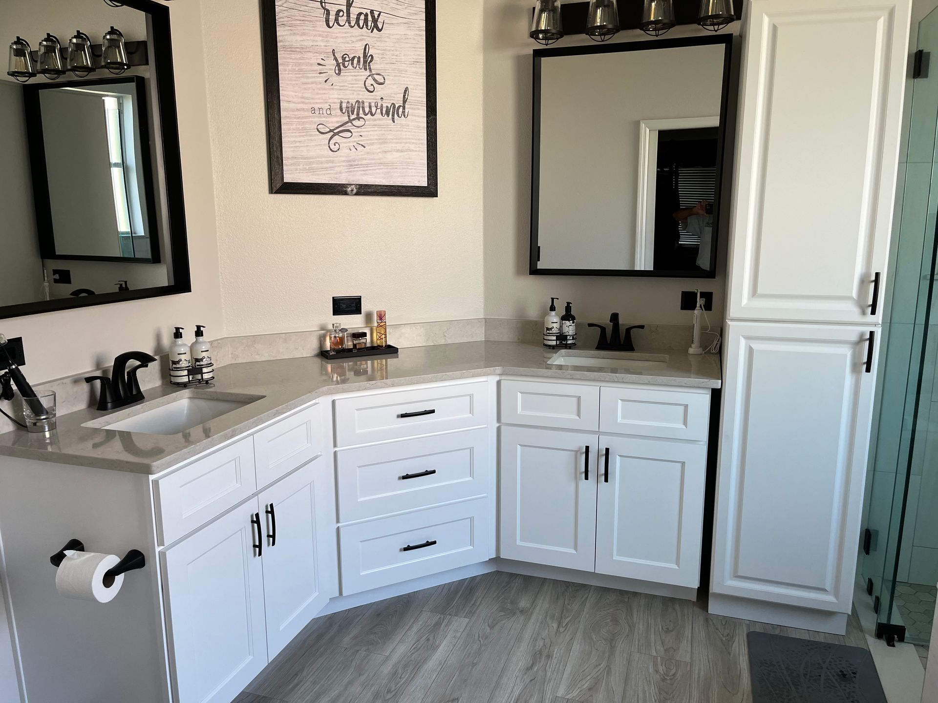 White bathroom with a corner vanity, two mirrors, and a tall cabinet. Gray countertop and light wood-look floor.