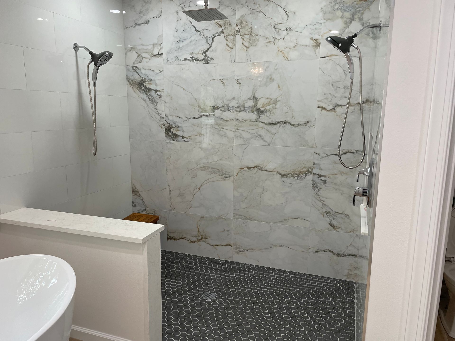 Modern bathroom with marble-look tiled shower. Features two shower heads, dark floor tile, and a white bench.
