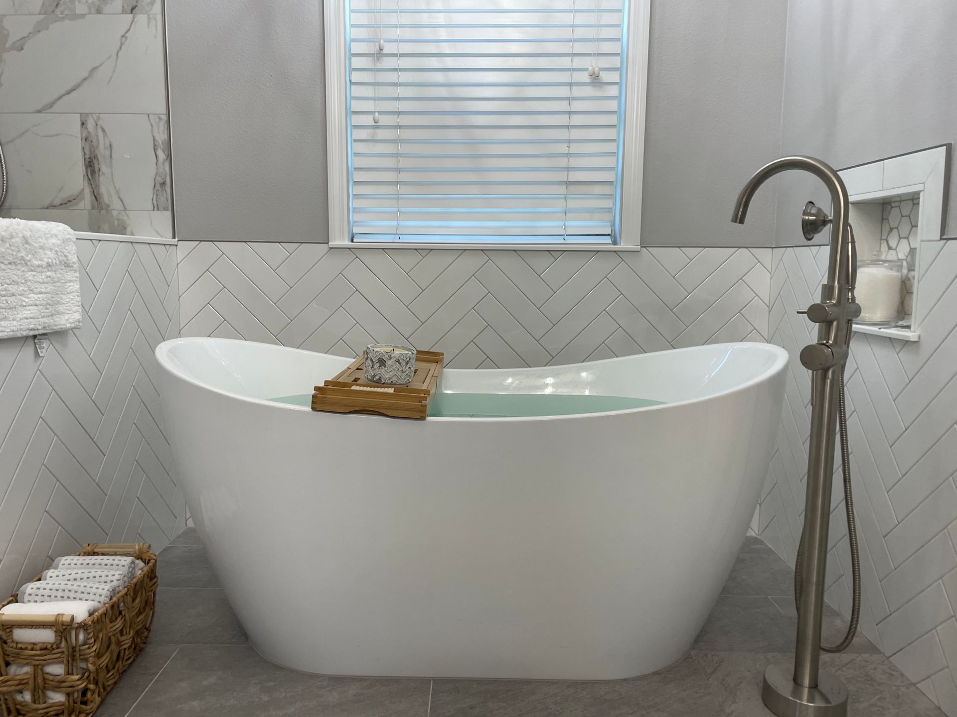 A white freestanding bathtub filled with water; a wooden tray holds soap. Gray and white tiled bathroom.