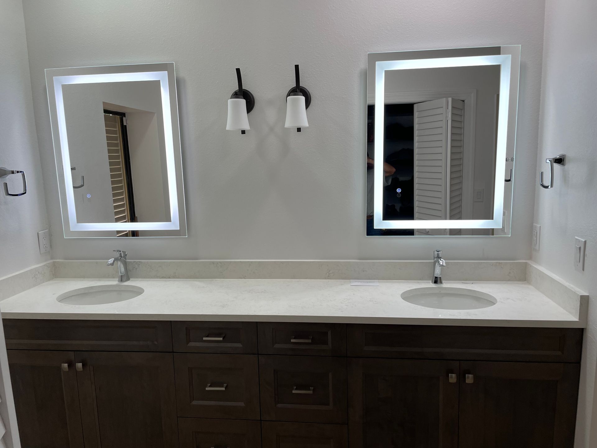 Bathroom vanity with two illuminated mirrors, sinks, brown cabinets, and sconces.