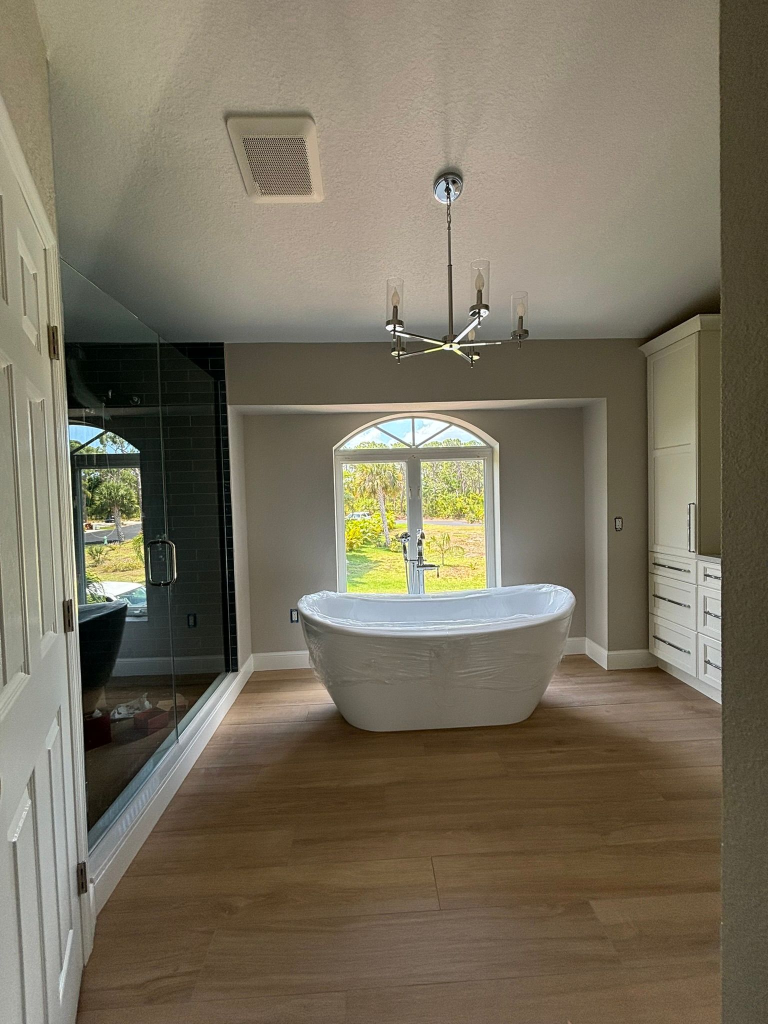 Bathroom with freestanding white tub under arched window, shower on left, wood-look floor, overhead light.