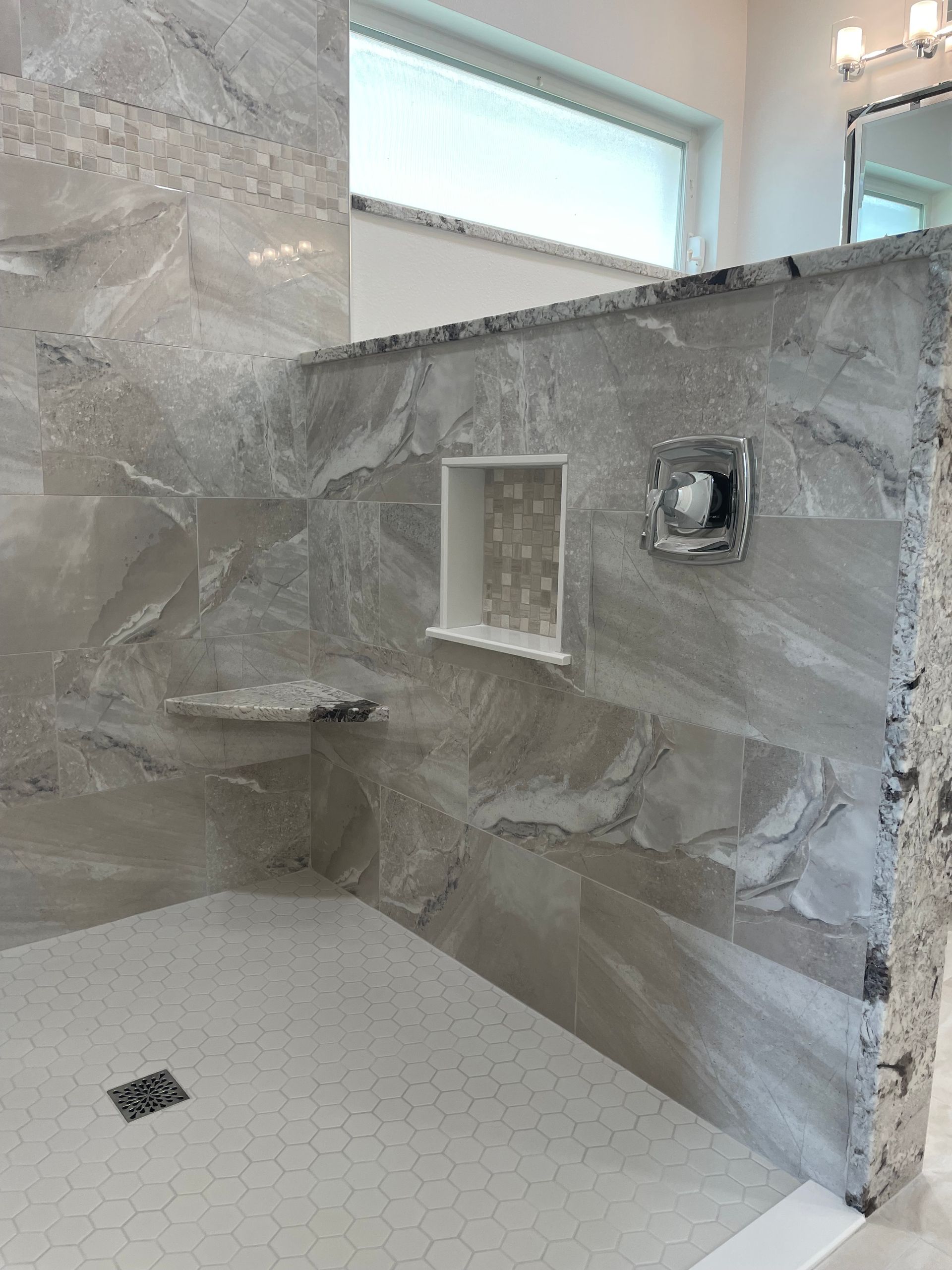 Shower stall with gray marbled tile walls, built-in shelf, recessed soap niche, and hexagonal floor tiles.