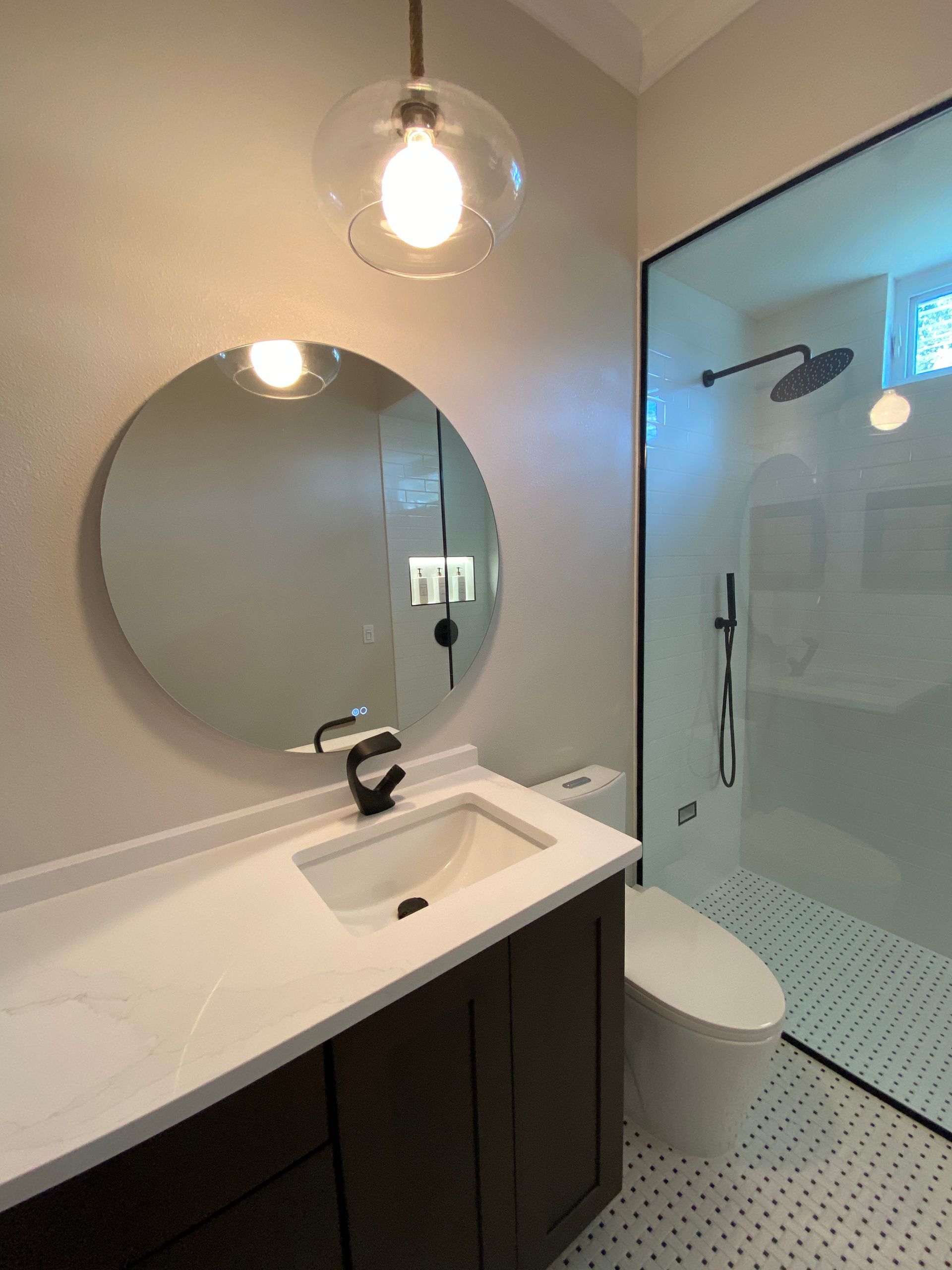 Modern bathroom with dark vanity, round mirror, glass-enclosed shower, and globe pendant light.