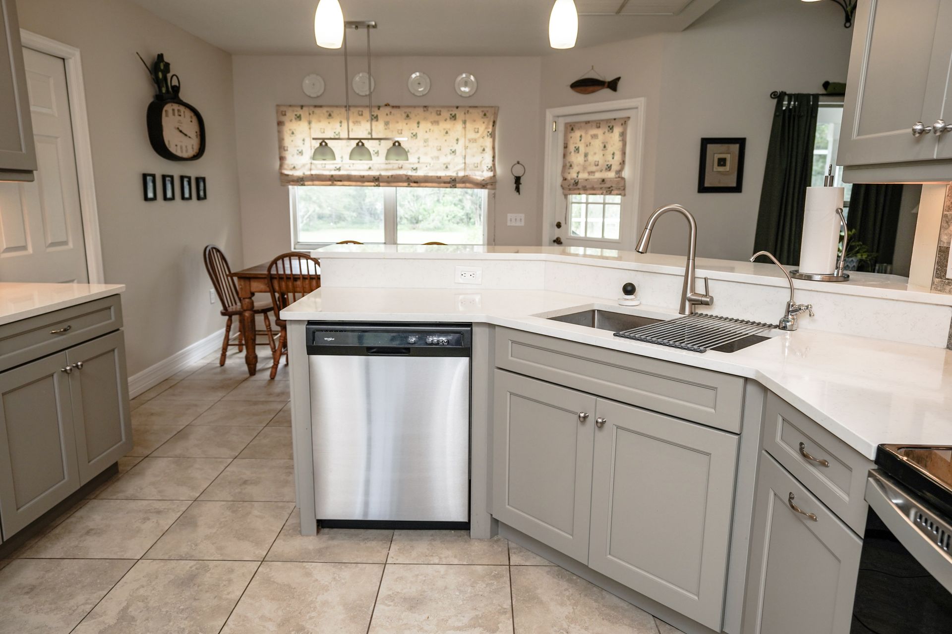 A kitchen with gray cabinets, white countertops, and stainless steel appliances. A window and dining table are in the background.